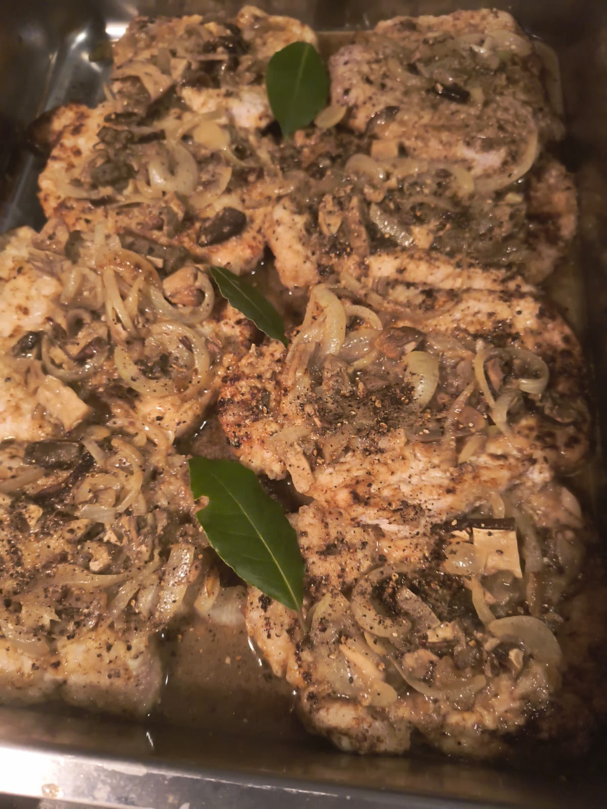 Pieczone plastry mięsa posypane pieprzem, z duszoną cebulą i grzybami, udekorowane liściem laurowym, ułożone w metalowej brytfannie.