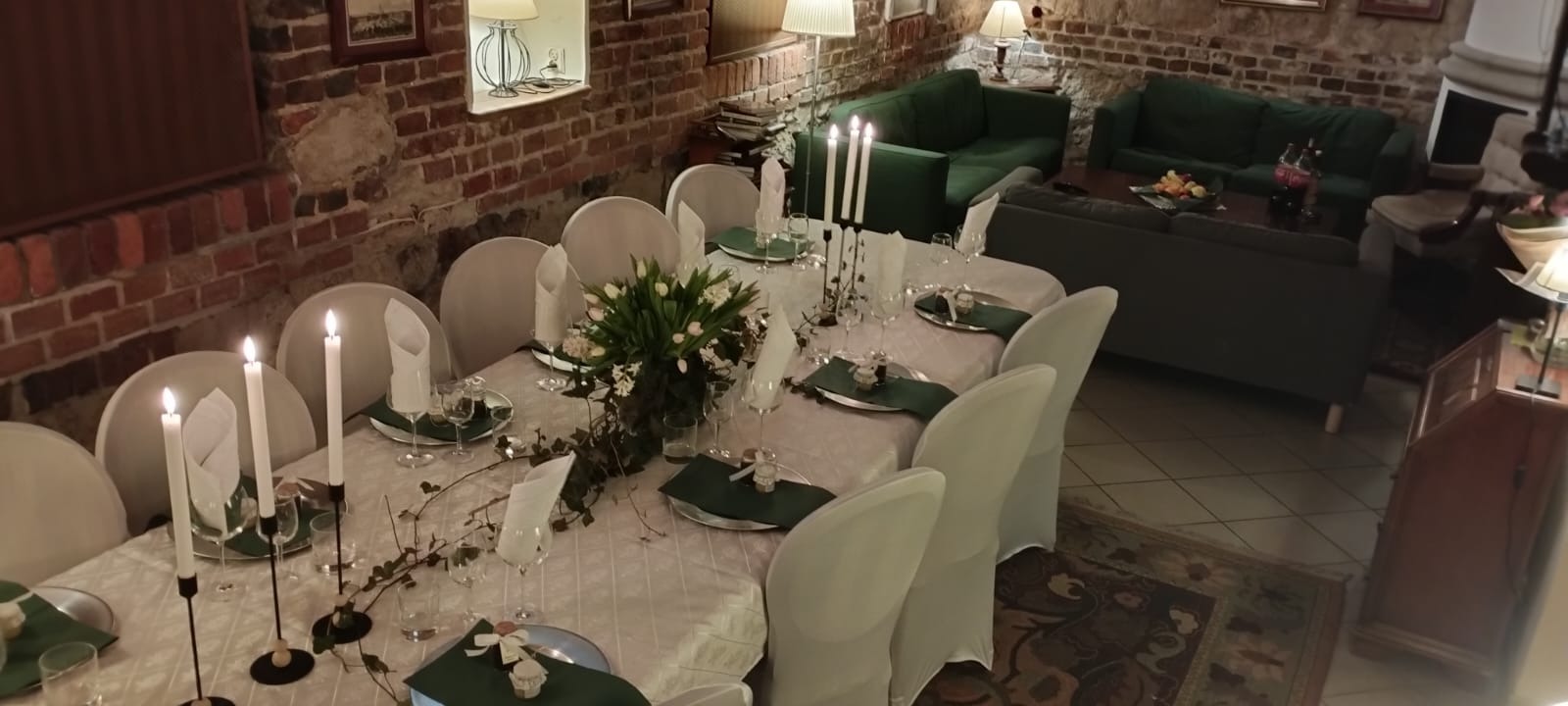 Uroczysty stół z białym obrusem, świecami, bukietem białych tulipanów i zielonymi serwetkami, przygotowany w pomieszczeniu z ceglanymi ścianami i zielonymi sofami w tle.