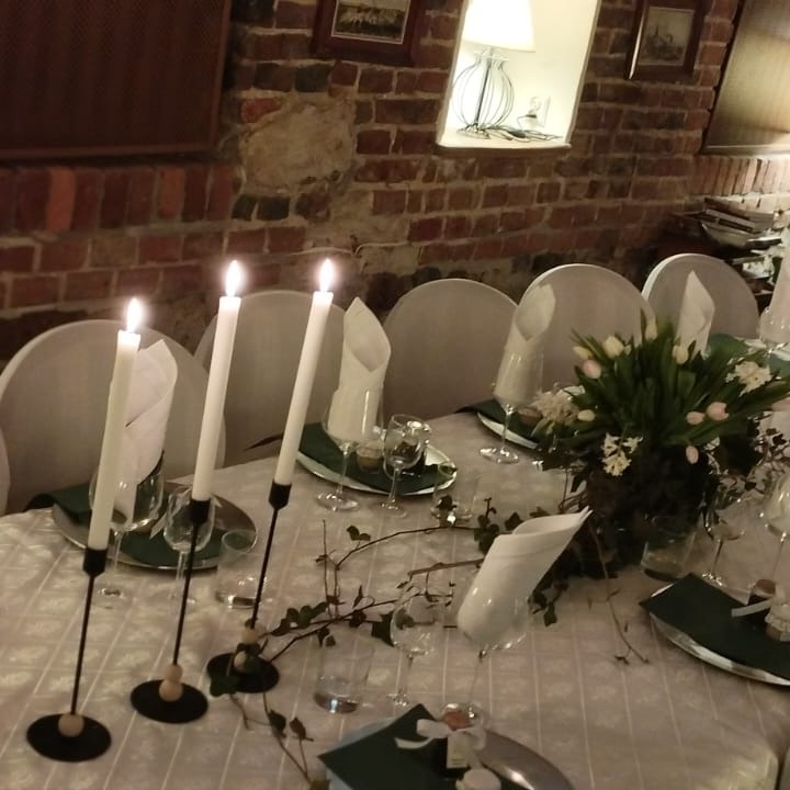 Uroczysty stół z białym obrusem, dekoracjami kwiatowymi, świecami w czarnych świecznikach i nakryciami w odcieniach bieli i zieleni, ustawiony w rustykalnym wnętrzu z ceglaną ścianą.