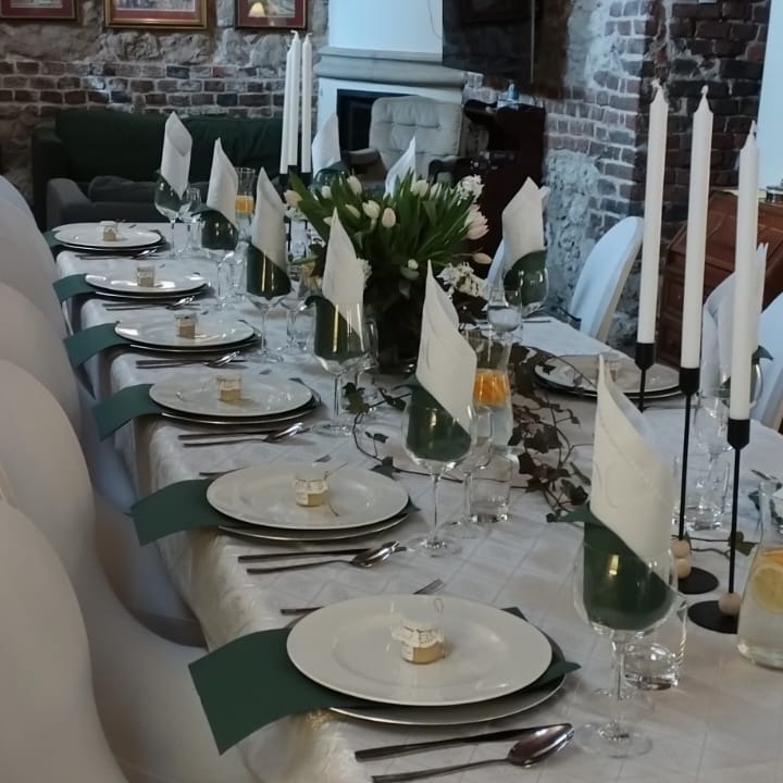 Uroczysty stół z białym obrusem w kratę, nakryty białymi talerzami z małymi upominkami, zielonymi serwetkami, kieliszkami z zielonymi wkładkami, wysokimi świecami i bukietem białych tulipanów...