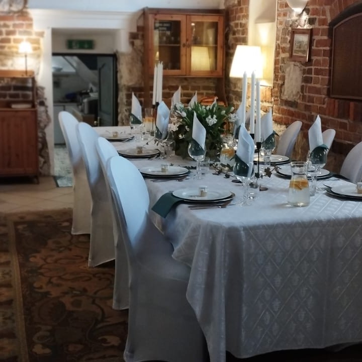 Uroczysty stół z białym obrusem, nakryty dla wielu gości, z białymi pokrowcami na krzesłach, dekorowany świecami, kwiatami i zielonymi serwetkami, ustawiony w pomieszczeniu z ceglanymi ścianami...