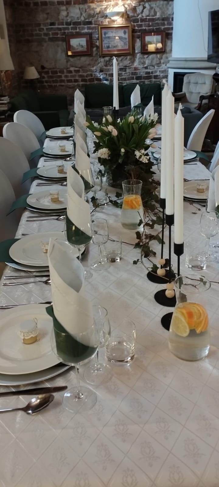 Uroczysty stół z białym obrusem w kwiatowy wzór, nakryty białymi talerzami, srebrnymi sztućcami, szklankami, kieliszkami oraz białymi serwetkami w zielonych pierścieniach; dekoracje stanowią...