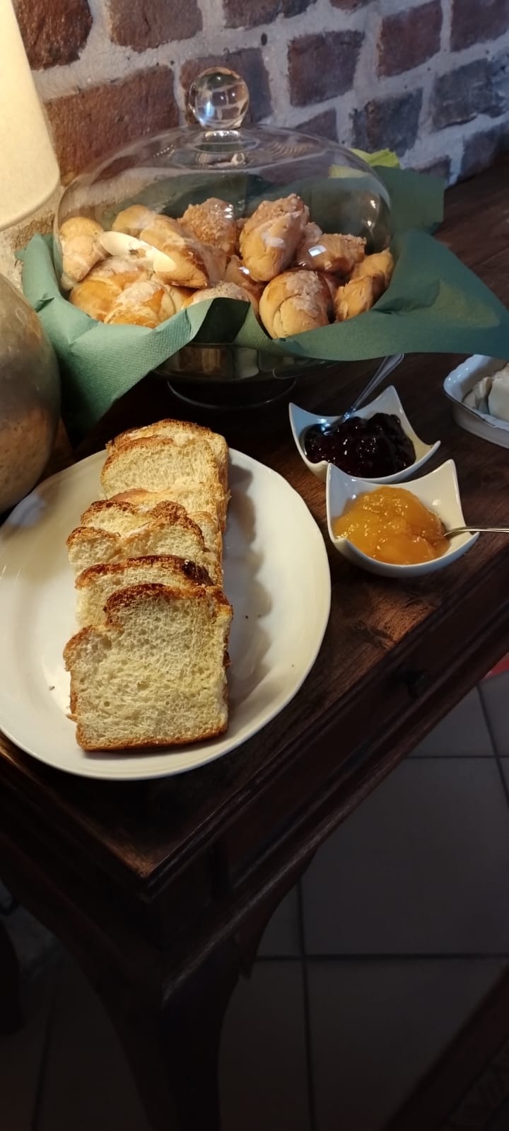 Śniadanie na ciemnym drewnianym stole: kromki chałki na białym talerzu, miseczki z dżemem pomarańczowym i ciemnym owocowym, koszyk z bułeczkami pod szklanym kloszem, ceglana ściana w tle.