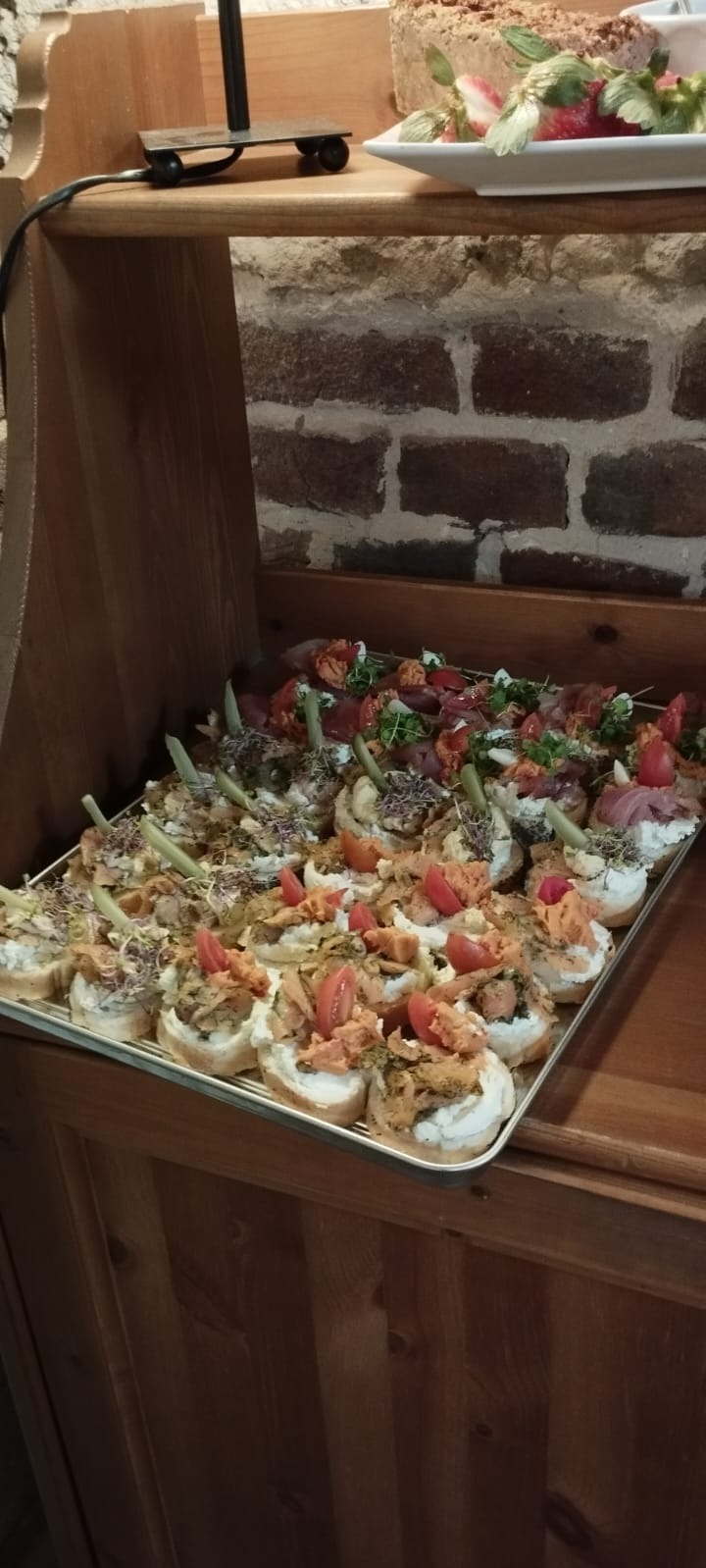 Taca z różnorodnymi kanapeczkami na białym pieczywie z dodatkami: wędlinami, pomidorami, kiełkami i warzywami, ustawiona na drewnianym regale z ceglanym tłem.