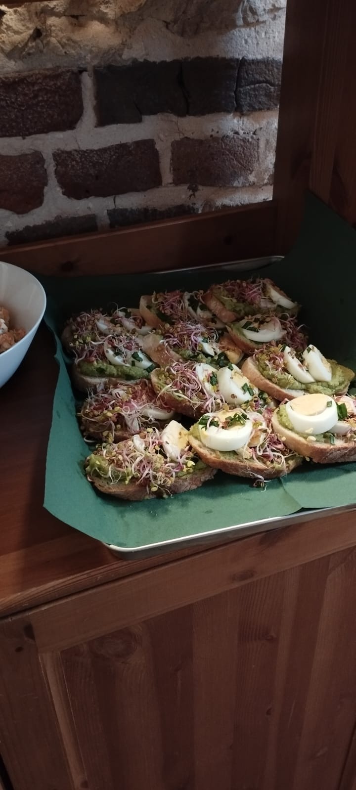 Taca kanapek z pastą z awokado, jajkiem na twardo i kiełkami, ułożonych na zielonym papierze, z ceglaną ścianą w tle.