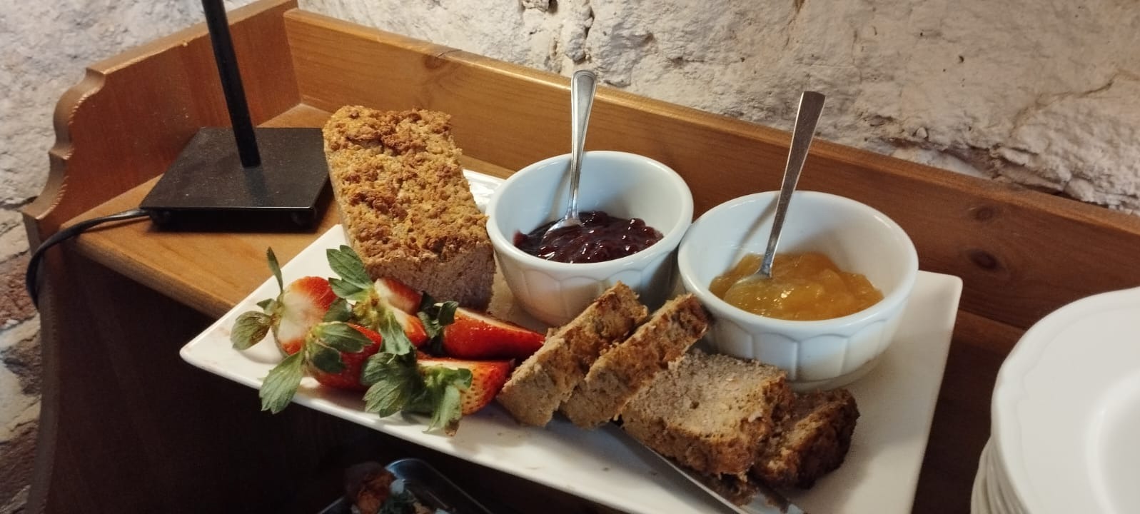 Deser na białym talerzu: keks z kruszonką, kromki pieczywa, truskawki i dwa białe naczynia z dżemem wiśniowym i brzoskwiniowym, każda z łyżeczką.