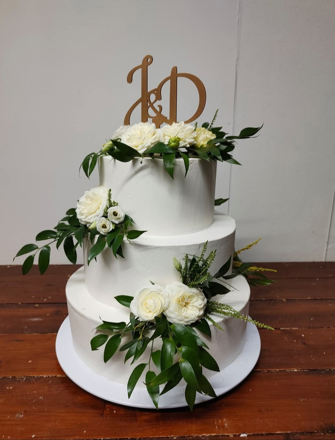 Trzypiętrowy tort weselny z białym lukrem, ozdobiony białymi różami i zielonymi liśćmi, zwieńczony drewnianym topperem z inicjałami 'J&D'.