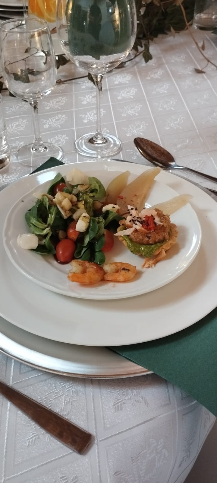 Elegancko podane danie na białym talerzu: sałatka z pomidorkami koktajlowymi i mini mozzarellą, grillowane krewetki oraz tarta z zielonym pesto i dekoracją z białych i czerwonych płatków na tle...