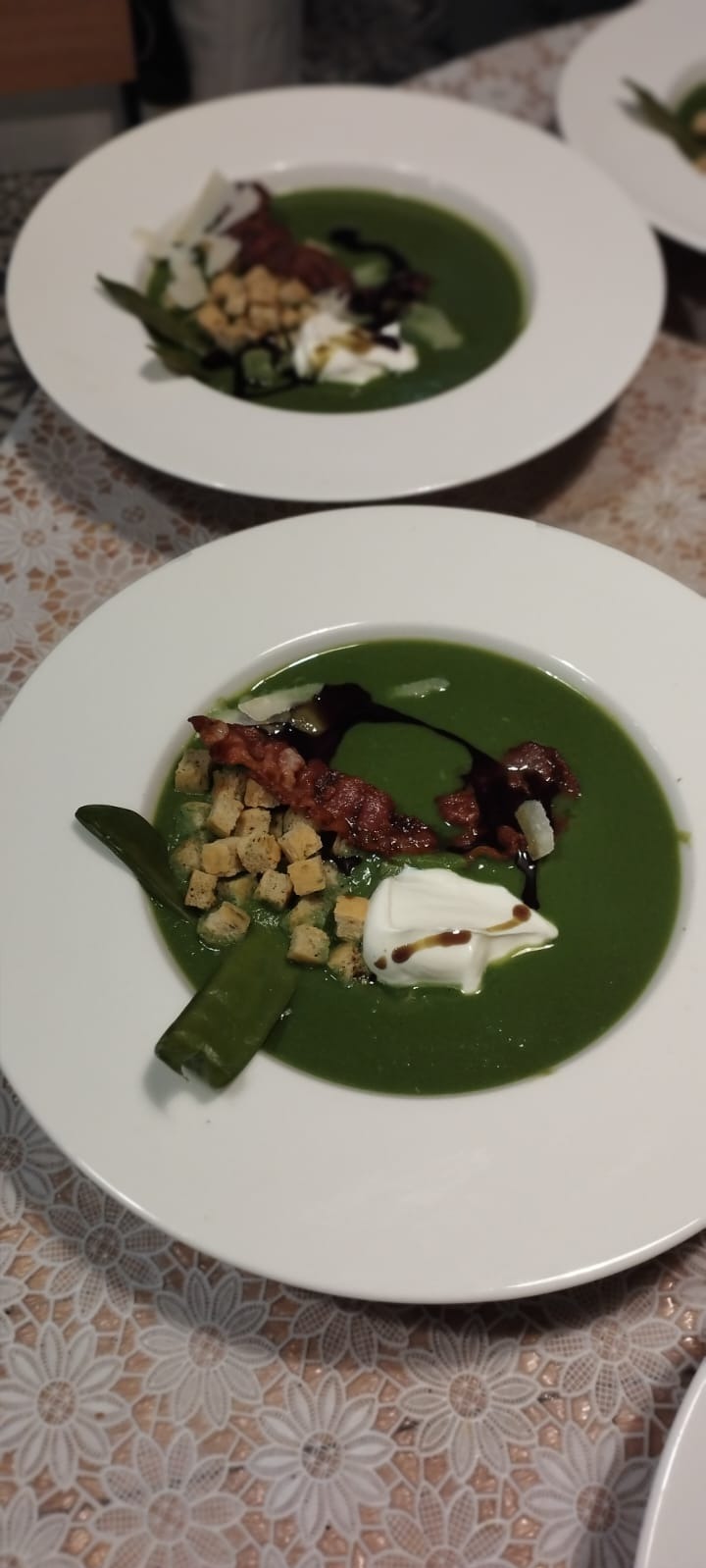 Porcja kremowej zielonej zupy na białym talerzu, udekorowana grzankami, bekonem, kleksem śmietany, sosem balsamicznym i zielonym strączkiem, na stole z koronkową serwetą.
