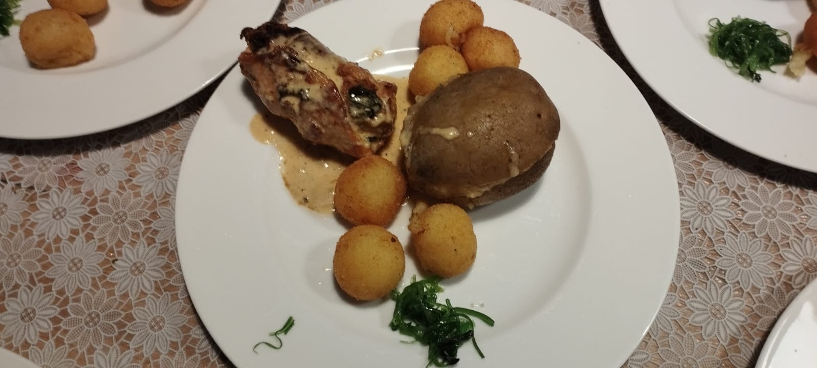 Elegancko podane danie na białym talerzu: pieczone mięso z sosem, ziemniak w mundurku i krokiety, udekorowane zielonymi algami, na stole z koronkowym obrusem.