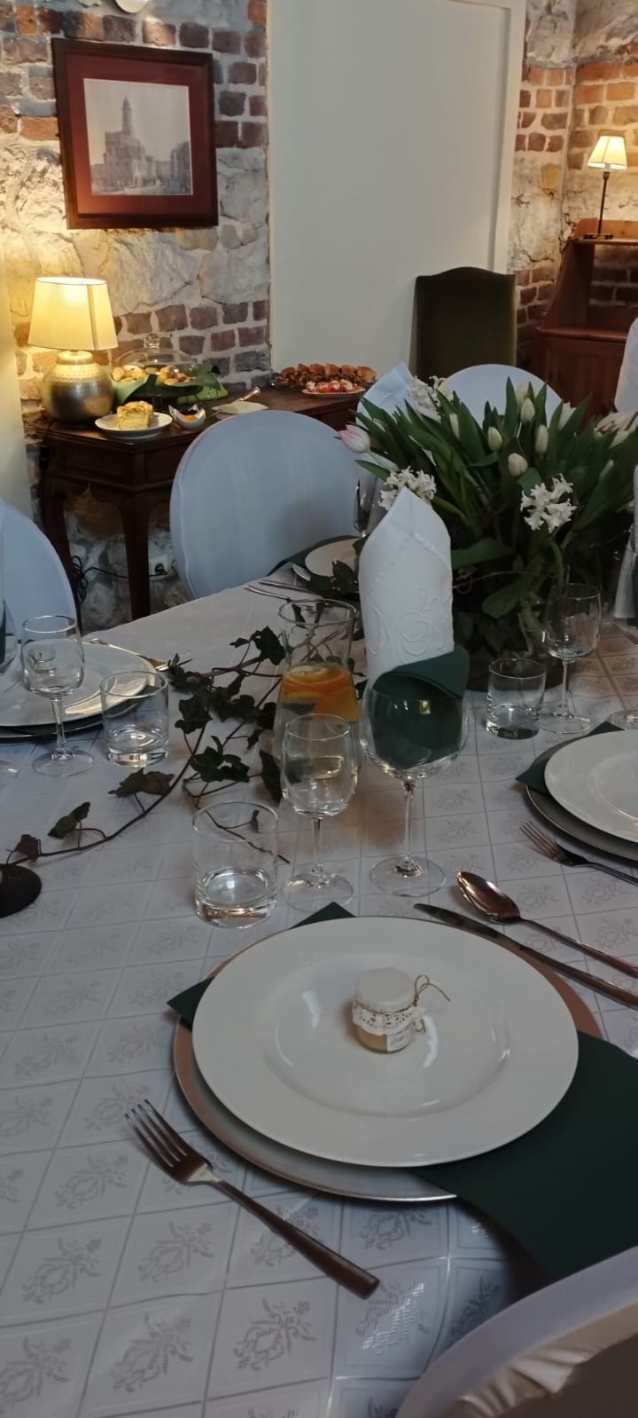Uroczysty stół z białym obrusem w delikatny wzór, elegancką zastawą, bukietem białych tulipanów i hiacyntów, szklankami z wodą i plastrem pomarańczy oraz małym słoiczkiem z miodem na każdym talerzu.