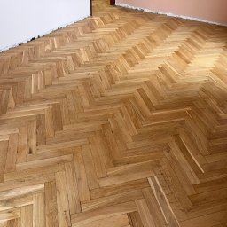 Pan Parkieciarz - Parkiet ułożony w jodełkę w jasnym odcieniu drewna, widoczne ślady po taśmie malarskiej na ścianie, grzejnik w rogu.