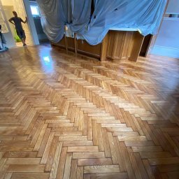 Pan Parkieciarz - Parkiet ułożony w jodełkę po cyklinowaniu, zabezpieczony folią mebel, widoczne maszyny do cyklinowania i pracownik w tle.