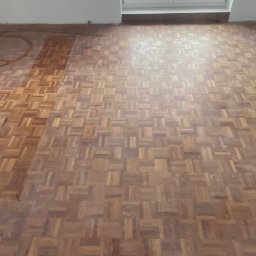 Pan Parkieciarz - Fragment drewnianej podłogi parkietowej w trakcie renowacji, z widoczną różnicą między obszarem po częściowym usunięciu starej powłoki a obszarem nietkniętym, na którym widoczne są ślady po okręgu.