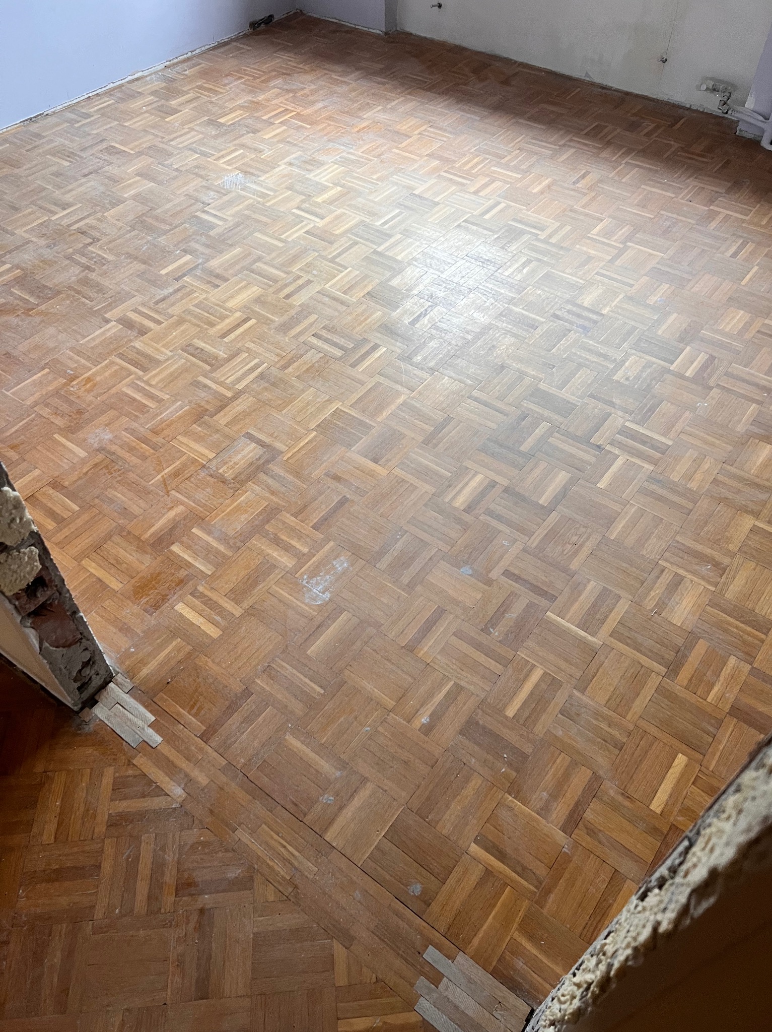 Parkiet ułożony w jodełkę na podłodze w pokoju, widoczne ślady użytkowania i drobne uszkodzenia, fragmenty uszkodzonej ściany i brakujących listew przypodłogowych.