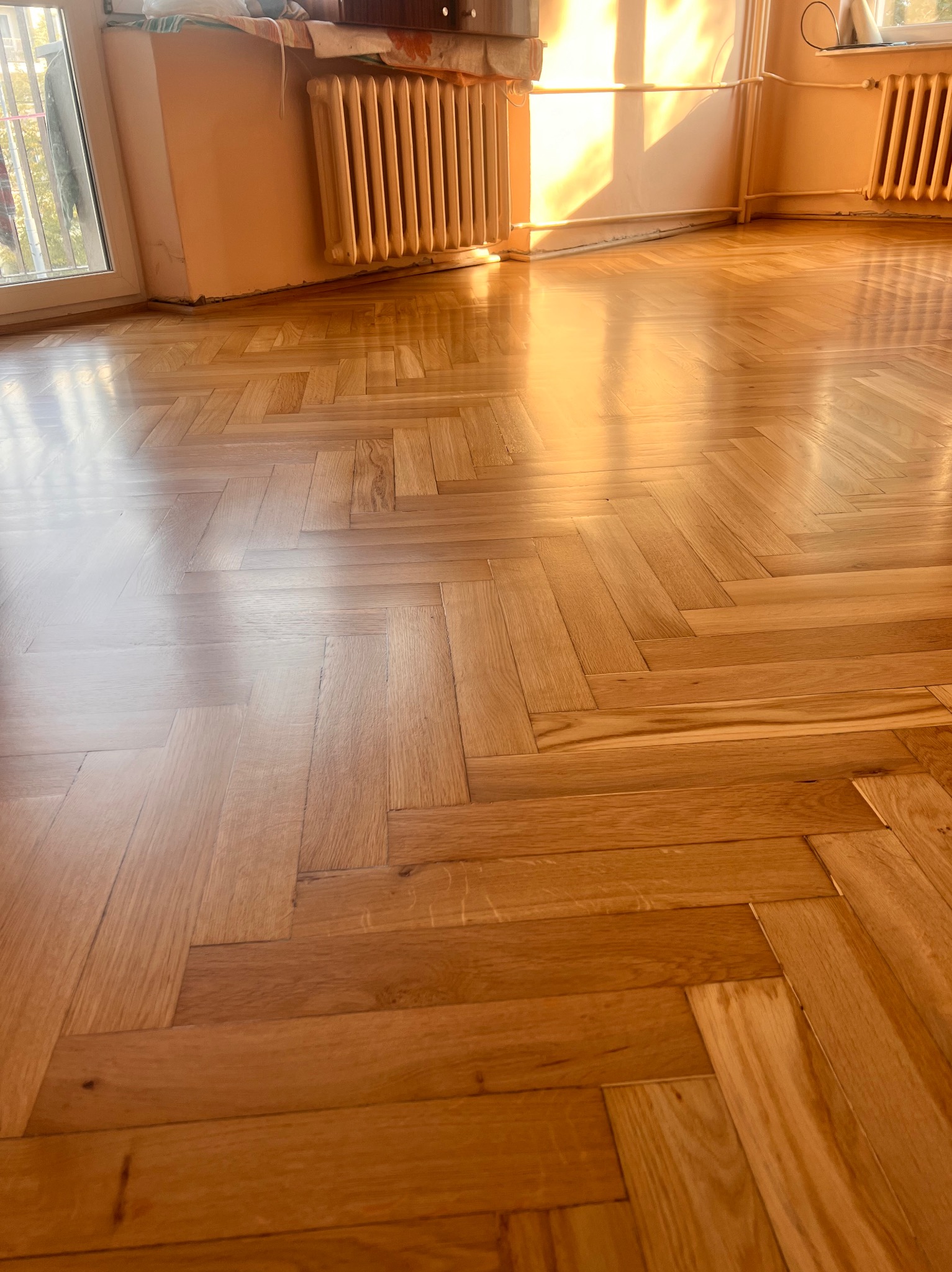 Parkiet ułożony w jodełkę, świeżo po renowacji, z widocznymi promieniami słońca wpadającymi przez okno, odbijającymi się od gładkiej powierzchni drewna i grzejnik w tle.