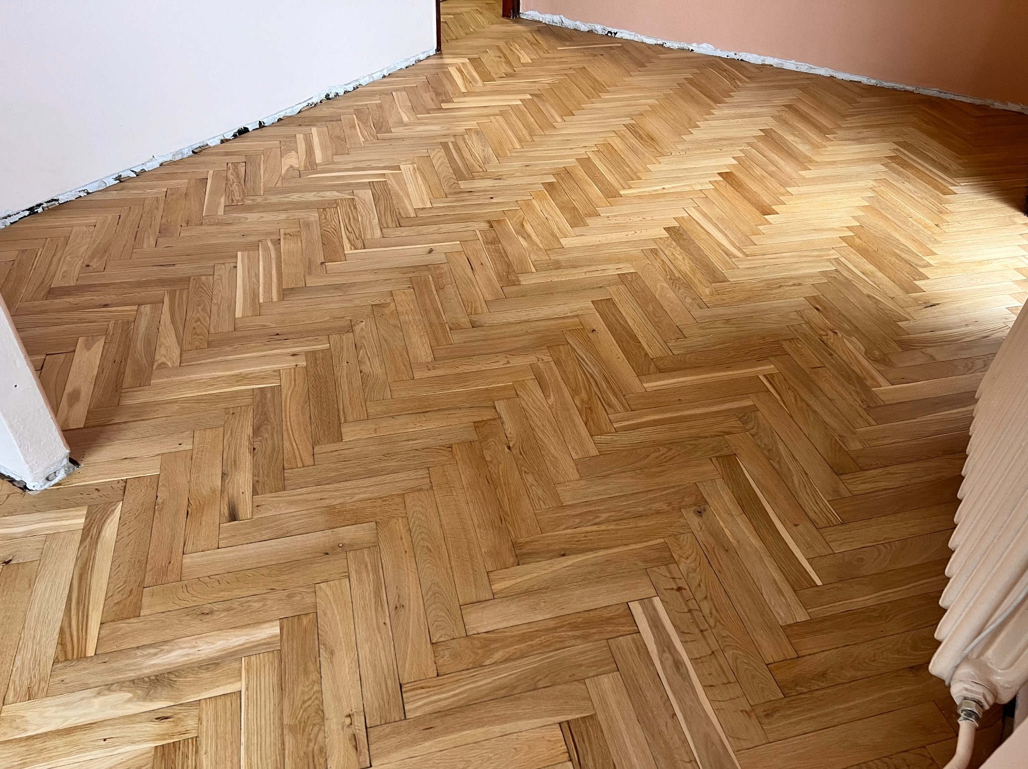 Parkiet ułożony w jodełkę w jasnym odcieniu drewna, widoczne ślady po taśmie malarskiej na ścianie, grzejnik w rogu.