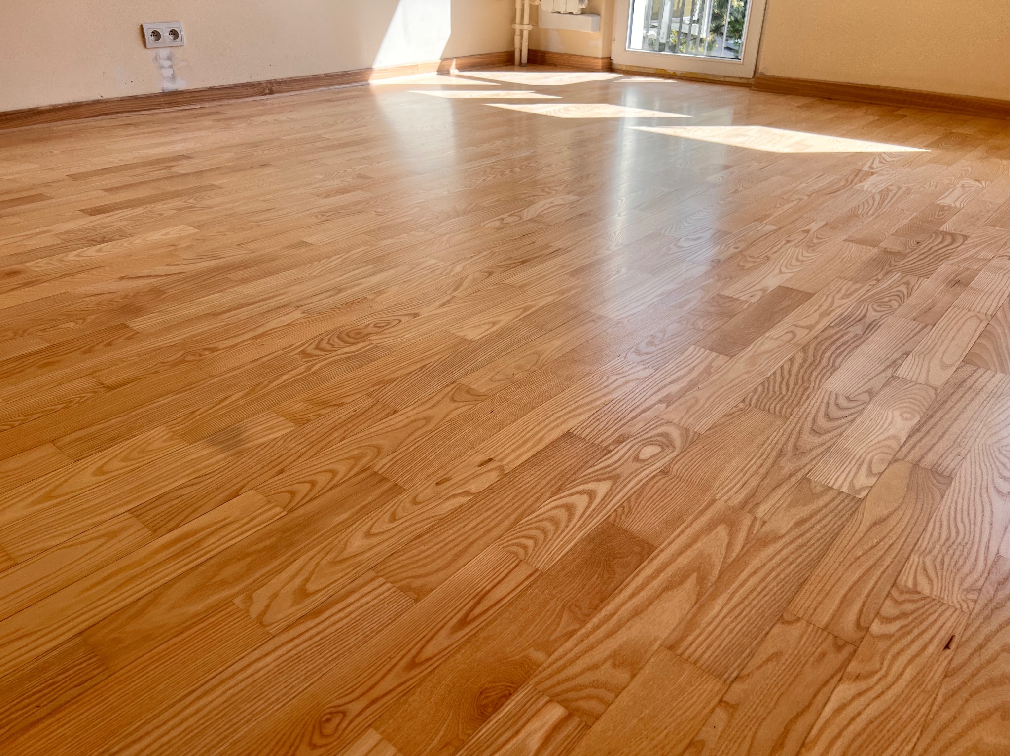 Wycyklinowana podłoga z jasnego drewna ułożona w jodełkę, widoczna ściana z gniazdkiem elektrycznym i listwa przypodłogowa, wpadające promienie słoneczne.