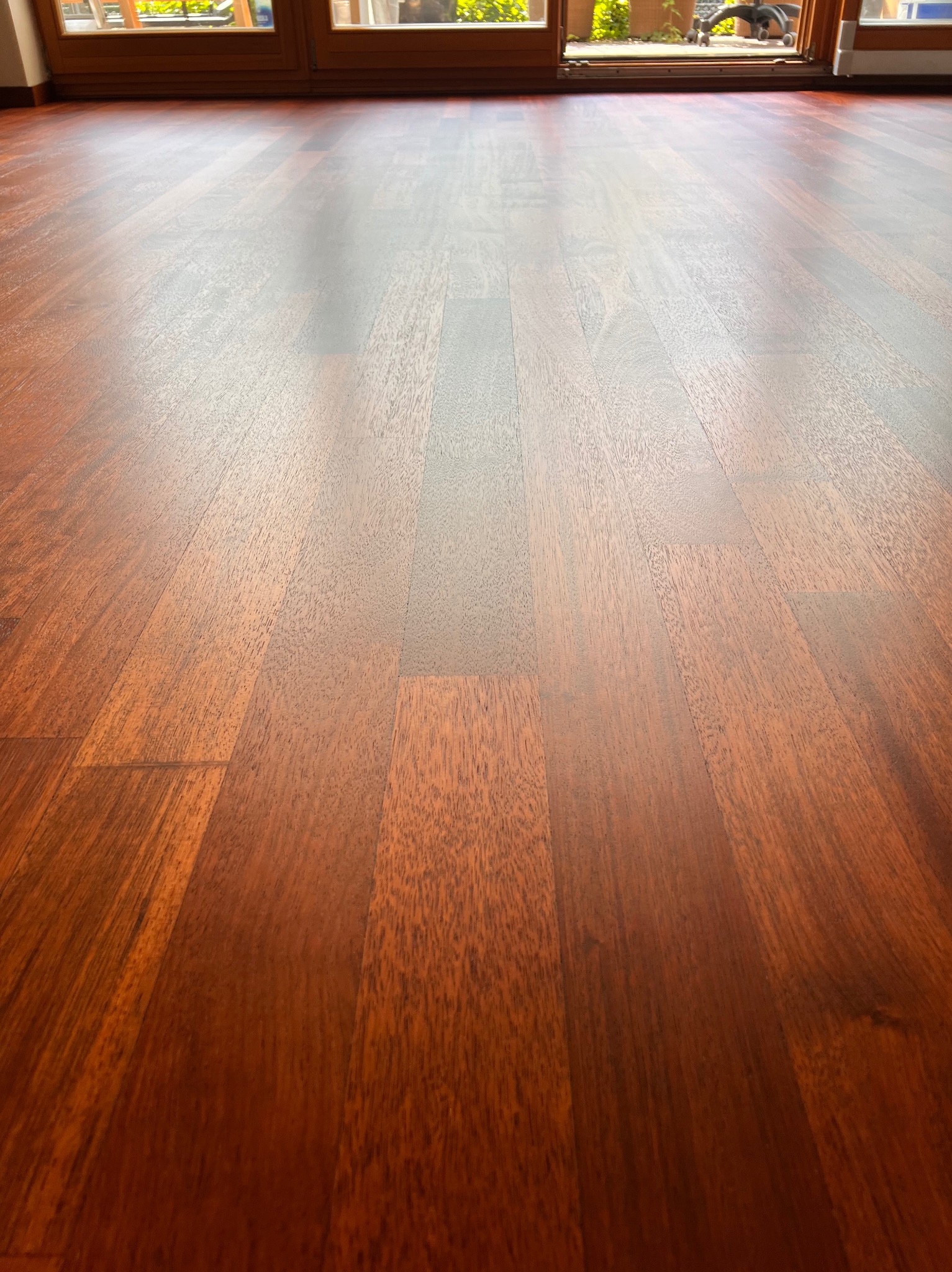 Błyszcząca, świeżo położona podłoga z ciemnego drewna egzotycznego, ułożona w parkiet, z widocznymi słojami i refleksem światła wpadającego przez przesuwne drzwi balkonowe.