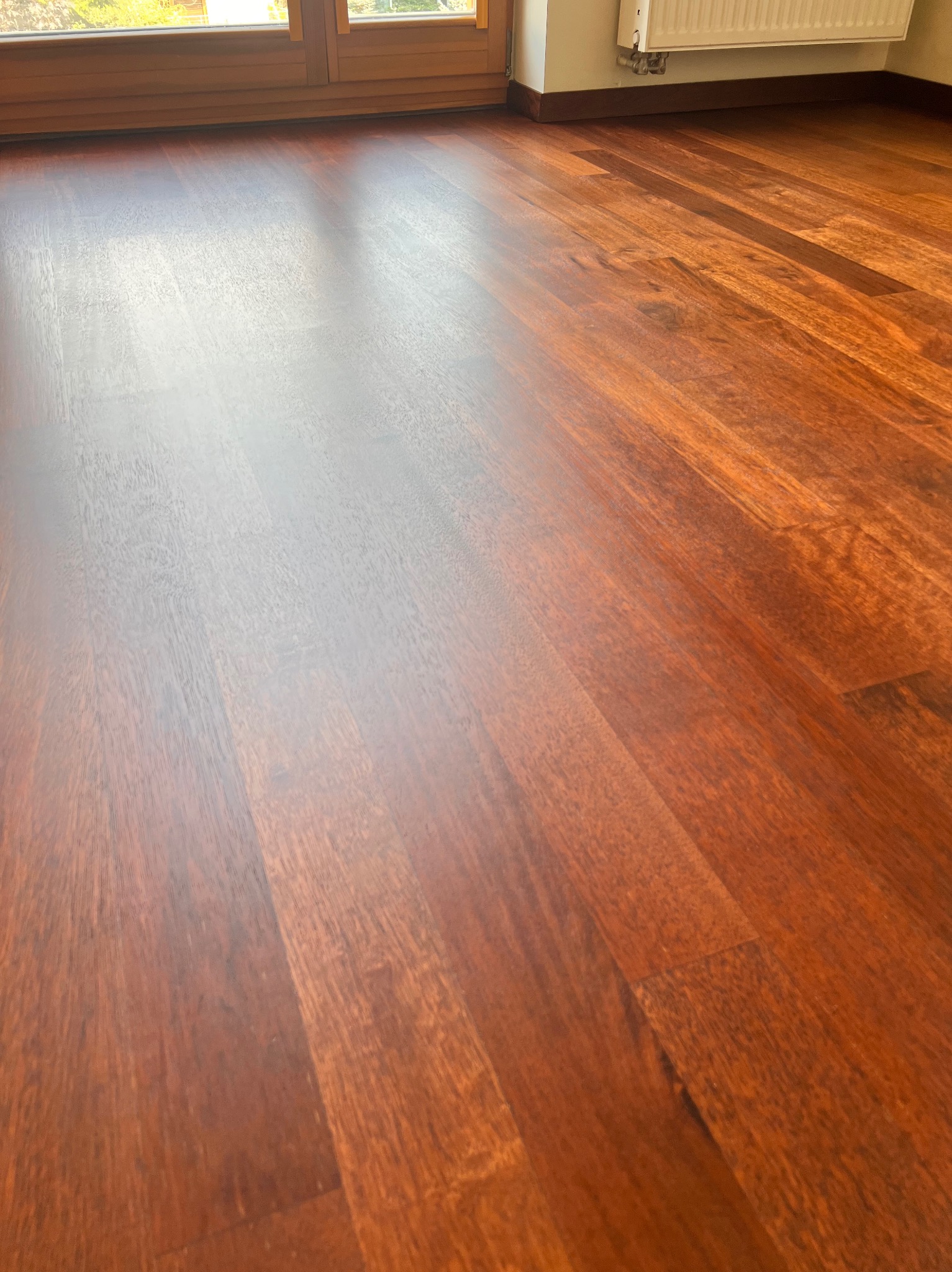 Polakierowana, brązowa podłoga z drewna egzotycznego, ułożona w deski, odbijająca światło słoneczne wpadające przez okno, z widocznym fragmentem grzejnika.