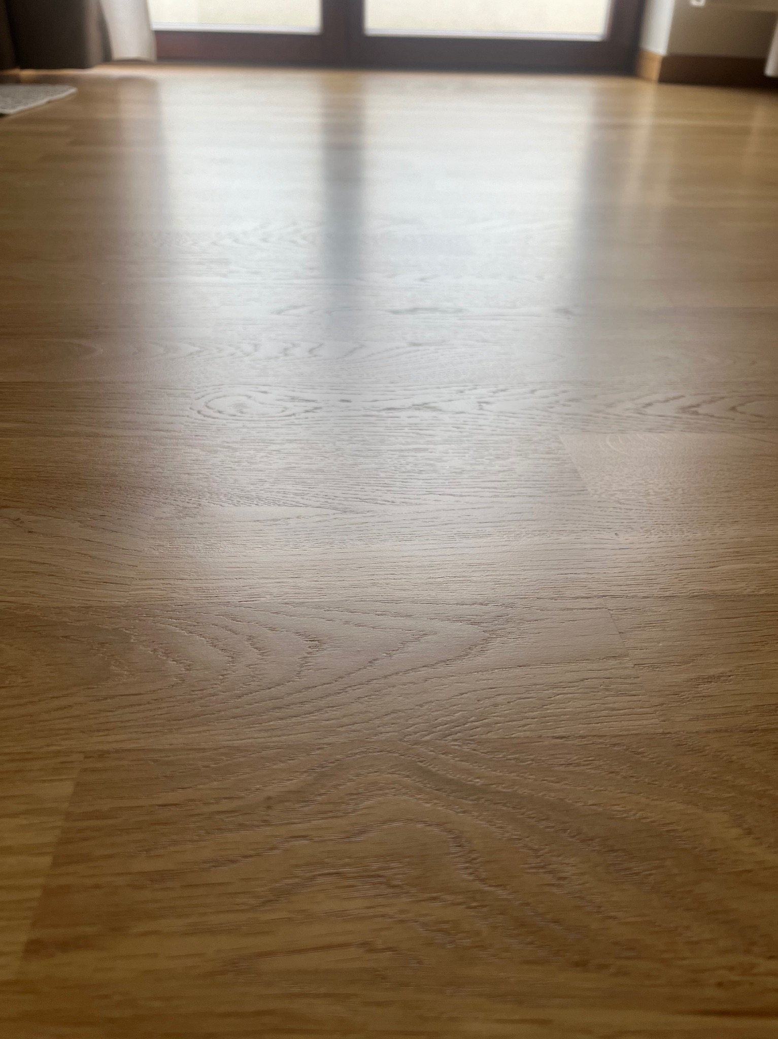 Jasne panele podłogowe z widocznym usłojeniem drewna, w tle rozmyte drzwi balkonowe.