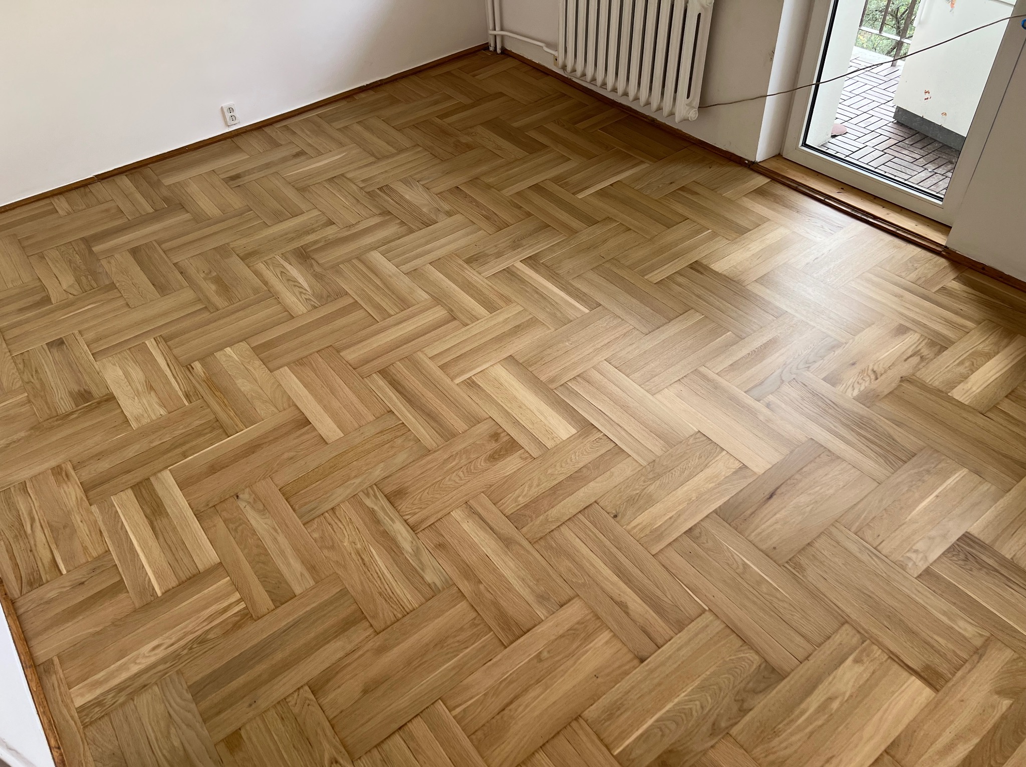 Parkiet ułożony w jodełkę francuską, po cyklinowaniu i lakierowaniu, widoczne detale usłojenia drewna, grzejnik i okno balkonowe w tle.