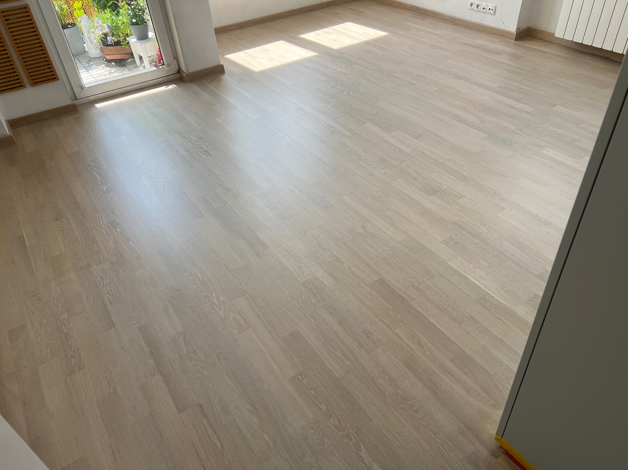 Jasne, odnowione panele podłogowe w pustym pokoju z widocznym fragmentem balkonu i promieniami słonecznymi wpadającymi przez okno.