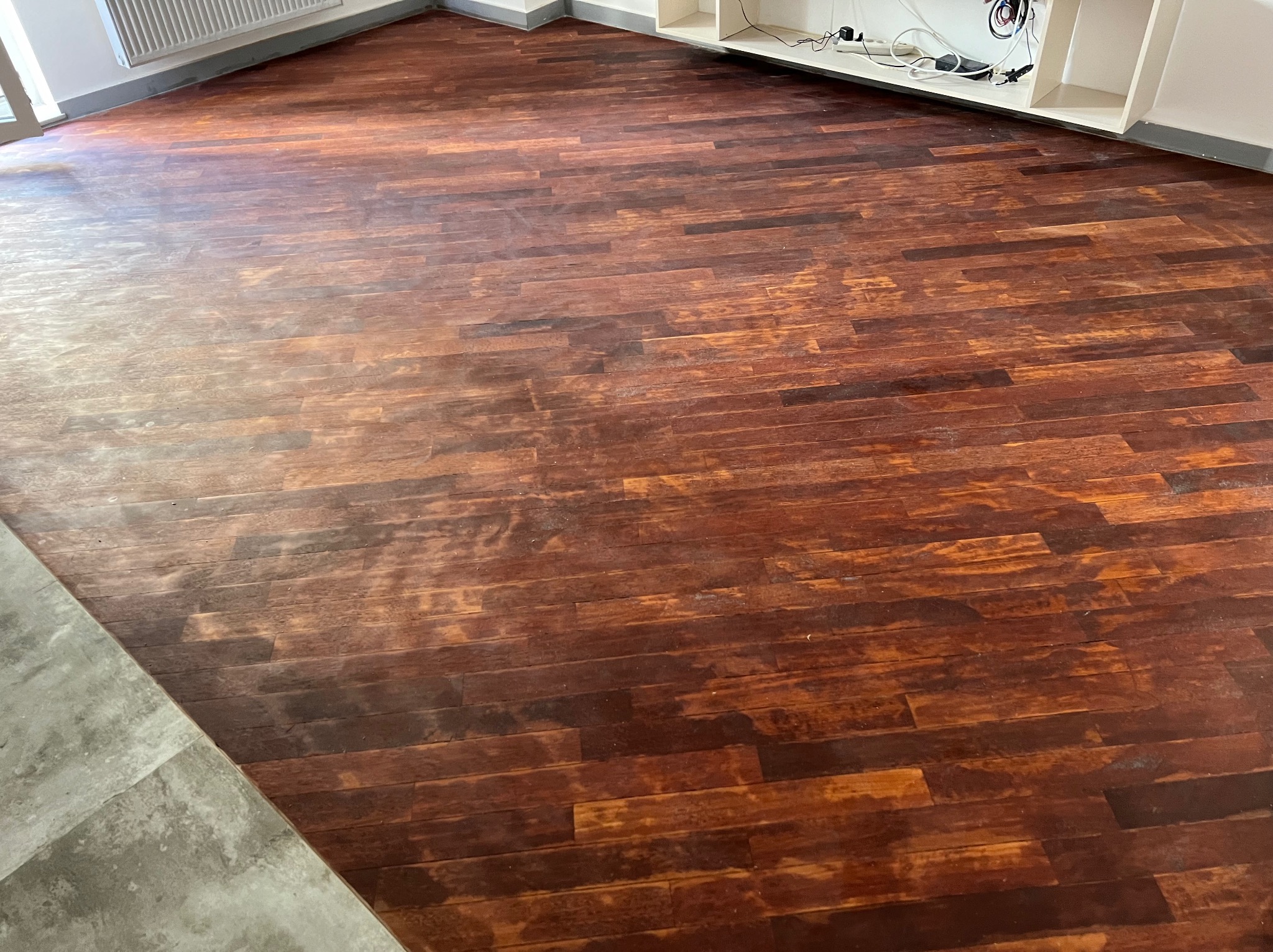 Ciemnobrązowa, odnowiona podłoga parkietowa z widocznymi śladami cyklinowania i lakierowania, z fragmentem szarej posadzki w rogu i białymi meblami w tle.