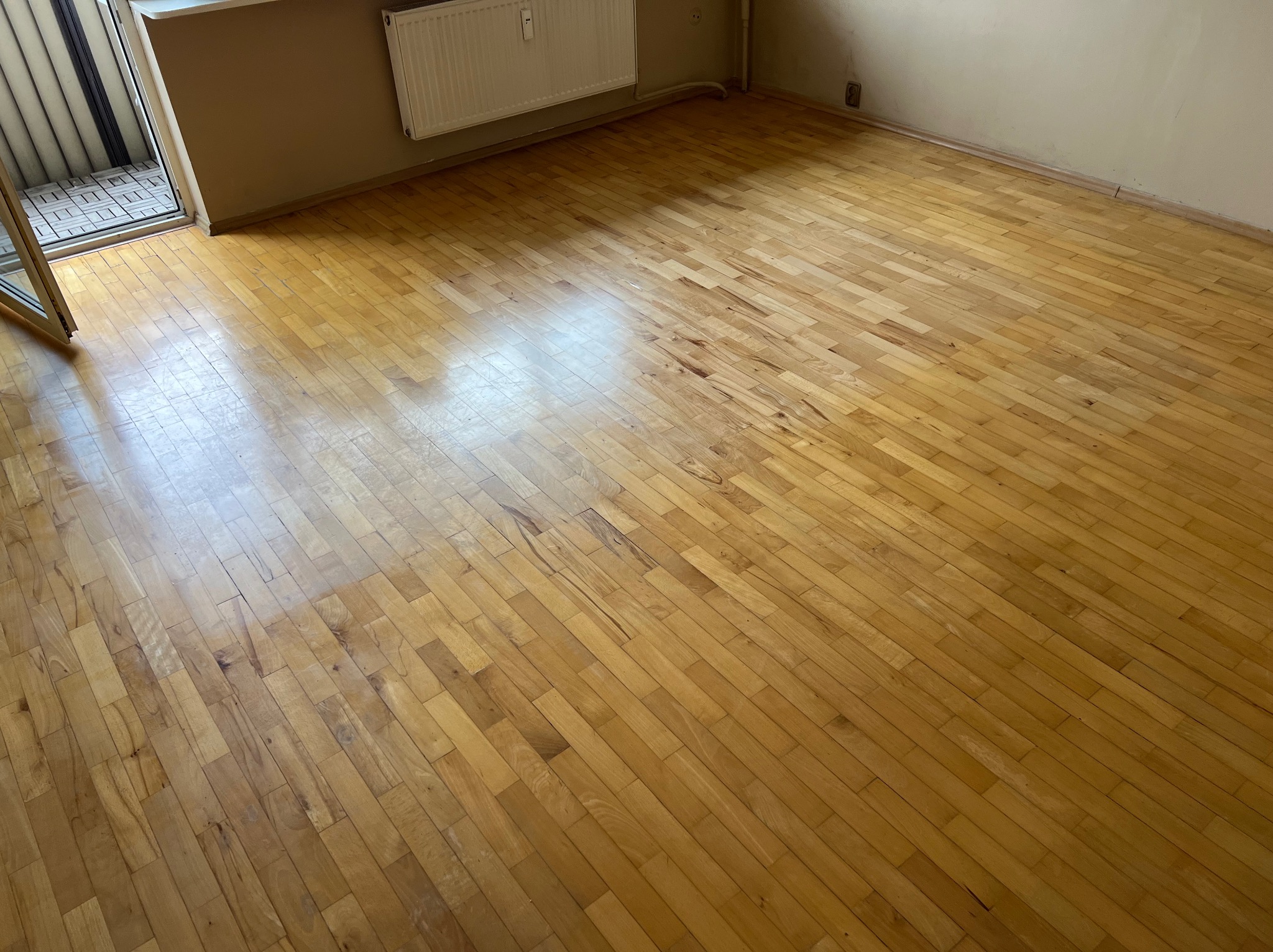 Jasna, drewniana podłoga parkietowa w pustym pokoju, widoczne ślady użytkowania i niewielkie ubytki w lakierze, z balkonem w tle i grzejnikiem na ścianie.