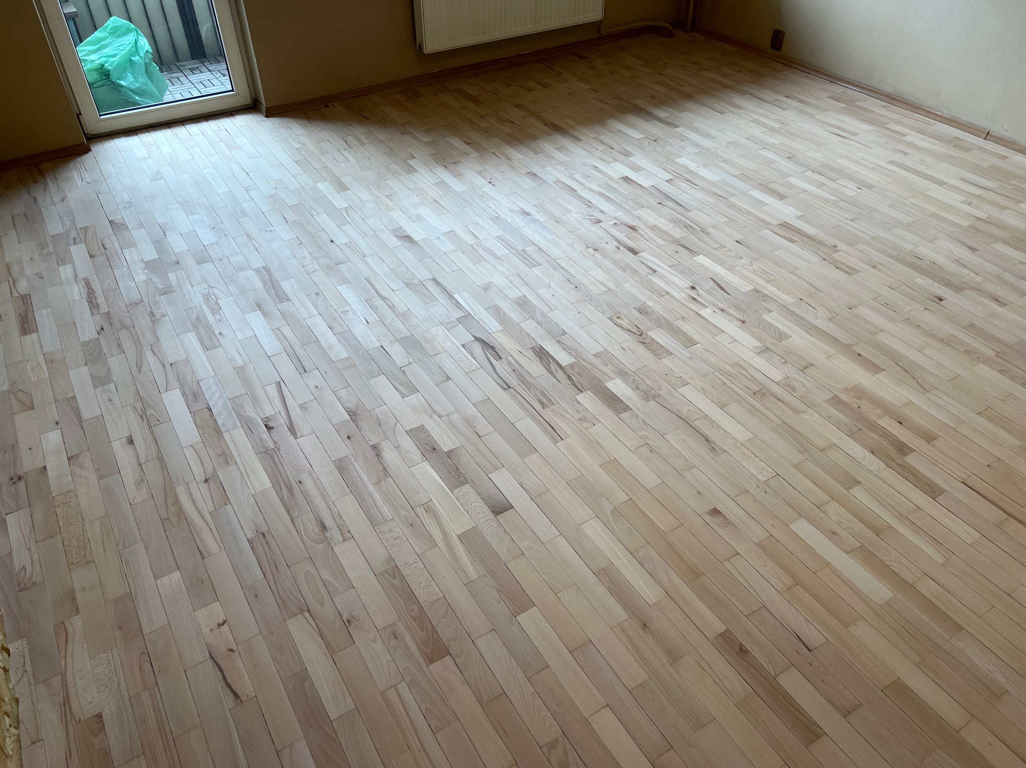 Jasna, nowo ułożona podłoga z parkietu w pustym pokoju, widoczne delikatne słoje drewna, w tle drzwi balkonowe z zielonym workiem.