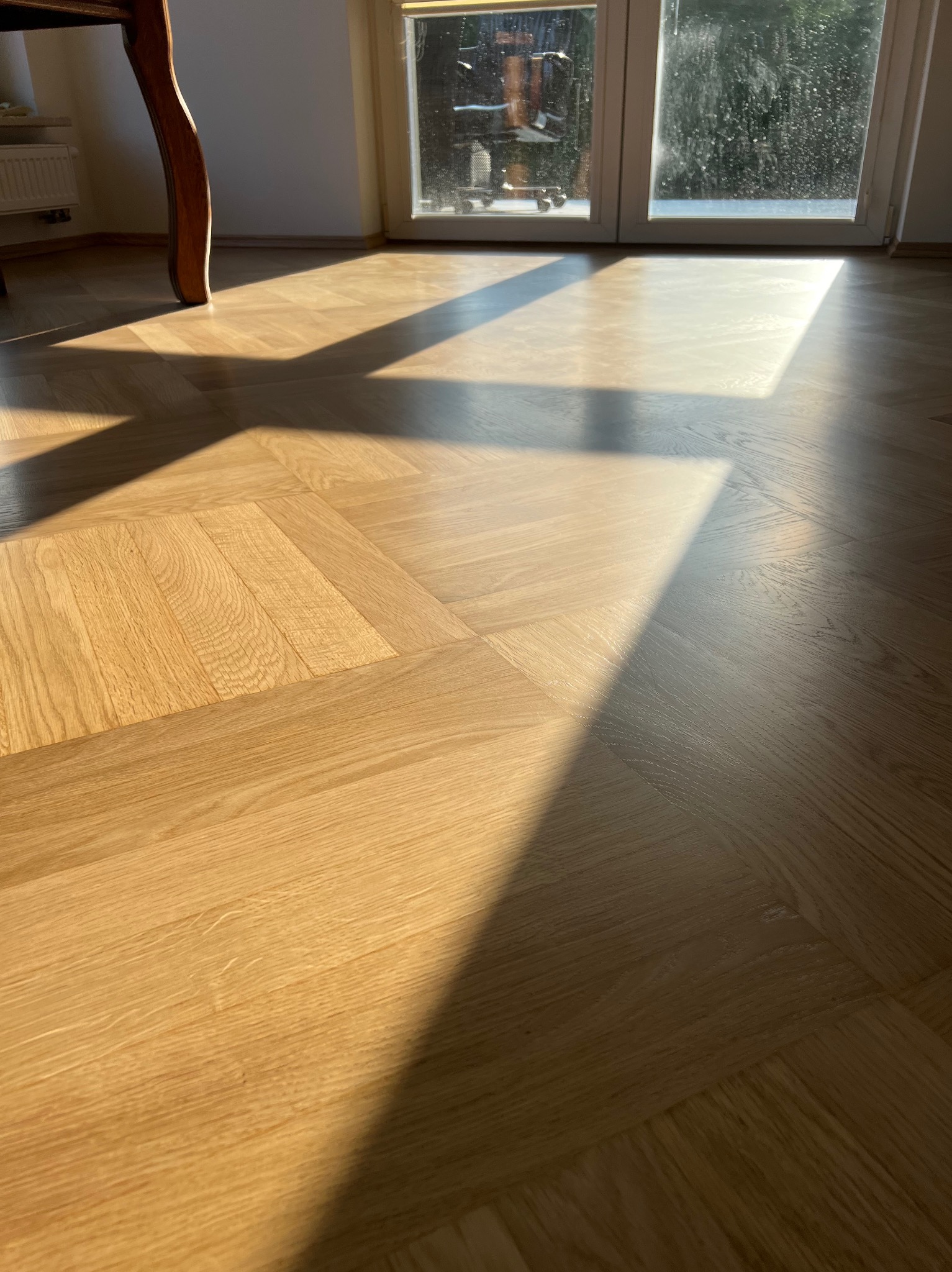 Parkiet ułożony we wzór jodełki, oświetlony promieniami słońca wpadającymi przez okno balkonowe, widoczny fragment nogi stołu.