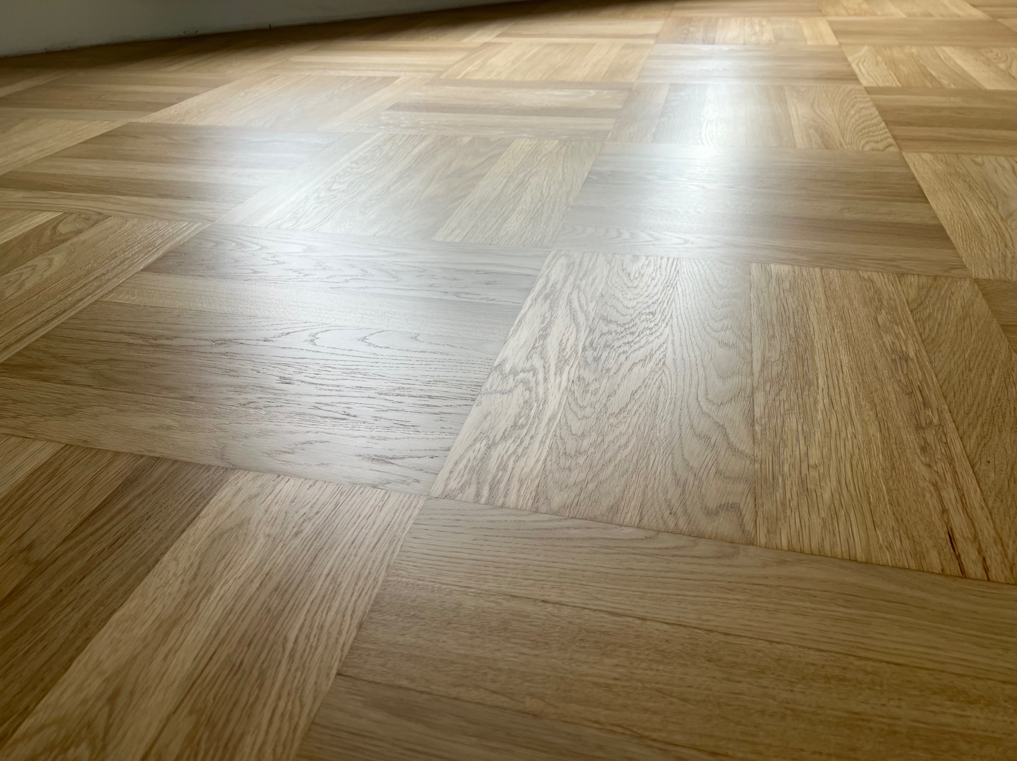 Parkiet ułożony w kwadraty z jasnego drewna dębowego, widoczne subtelne różnice w odcieniach poszczególnych klepek, z delikatnym połyskiem lakieru odbijającym światło.