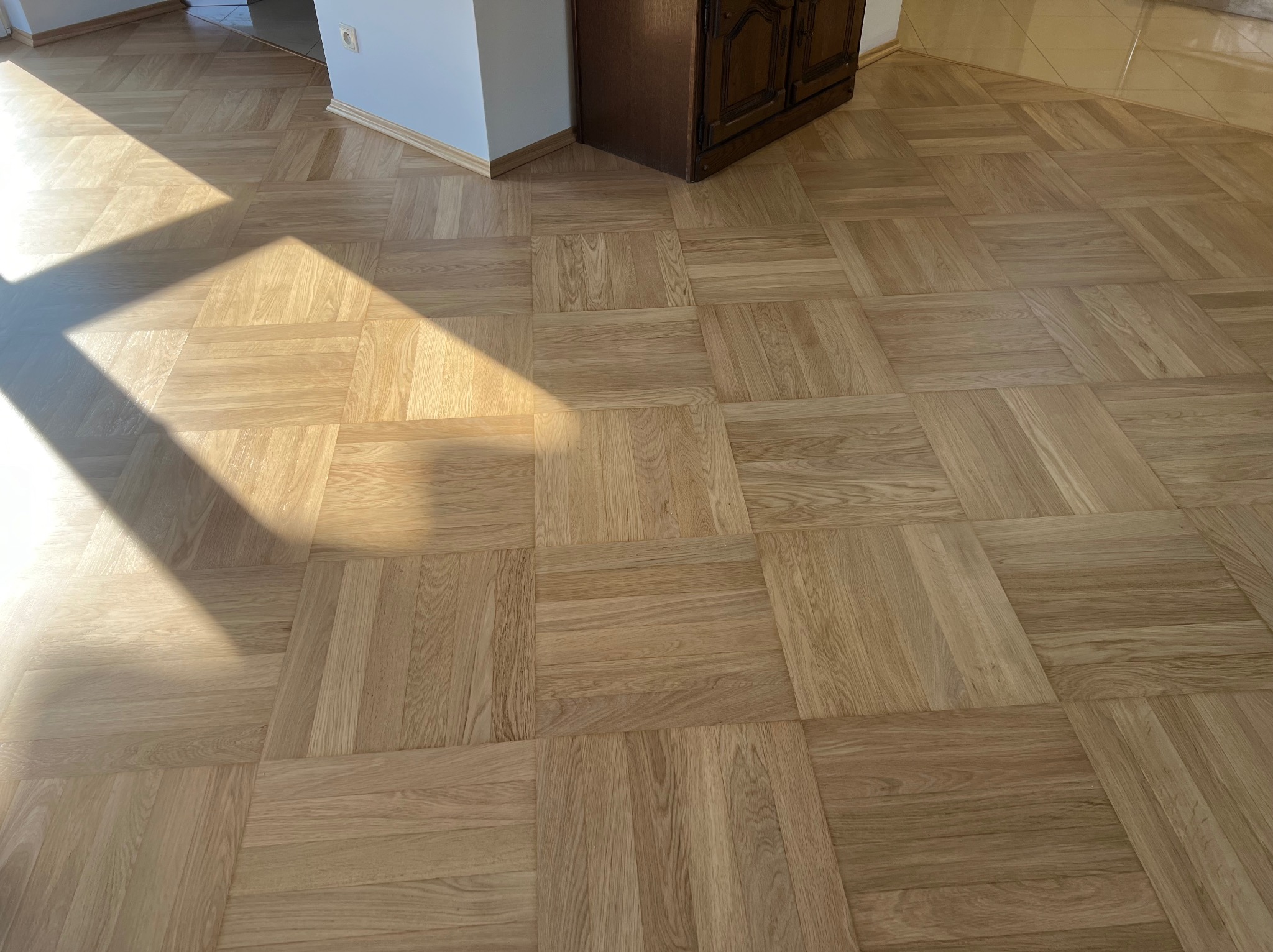 Parkiet ułożony w kwadraty z naturalnego drewna dębowego, oświetlony popołudniowym słońcem wpadającym przez okno, widoczny fragment brązowej szafki i biała ściana z listwą przypodłogową.