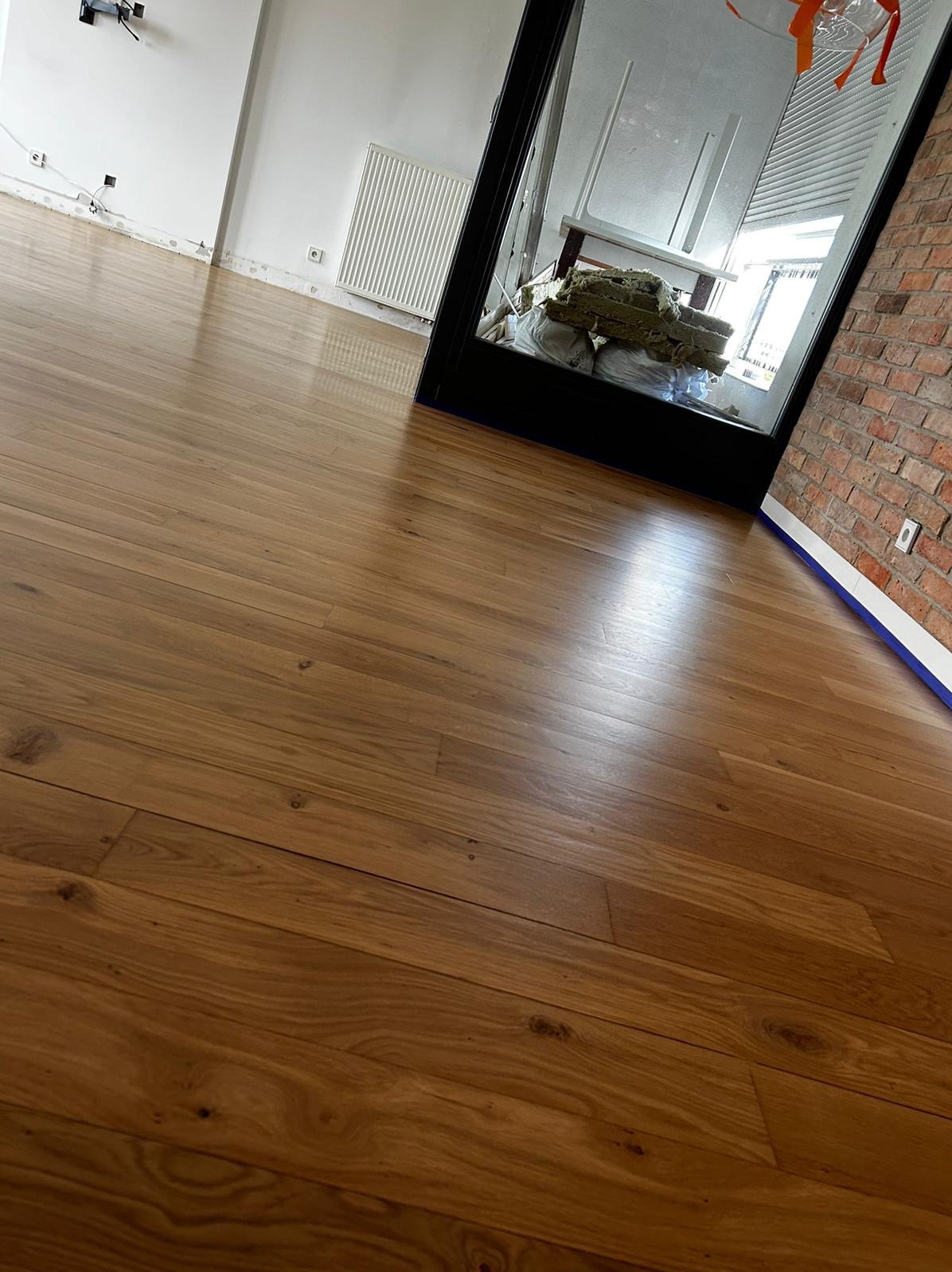 Polakierowana, drewniana podłoga z widocznymi słojami, świeżo po ułożeniu, z fragmentem białej ściany, grzejnikiem i ceglaną ścianą w tle.