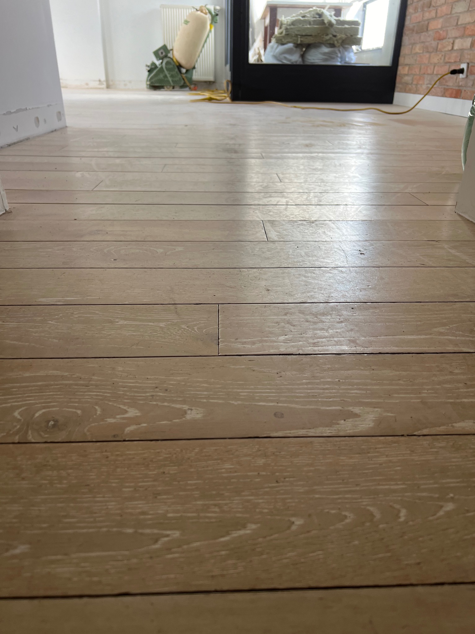 Jasne, drewniane deski podłogowe po częściowym wyszlifowaniu, z maszyną do cyklinowania w tle, widoczne ślady pyłu.