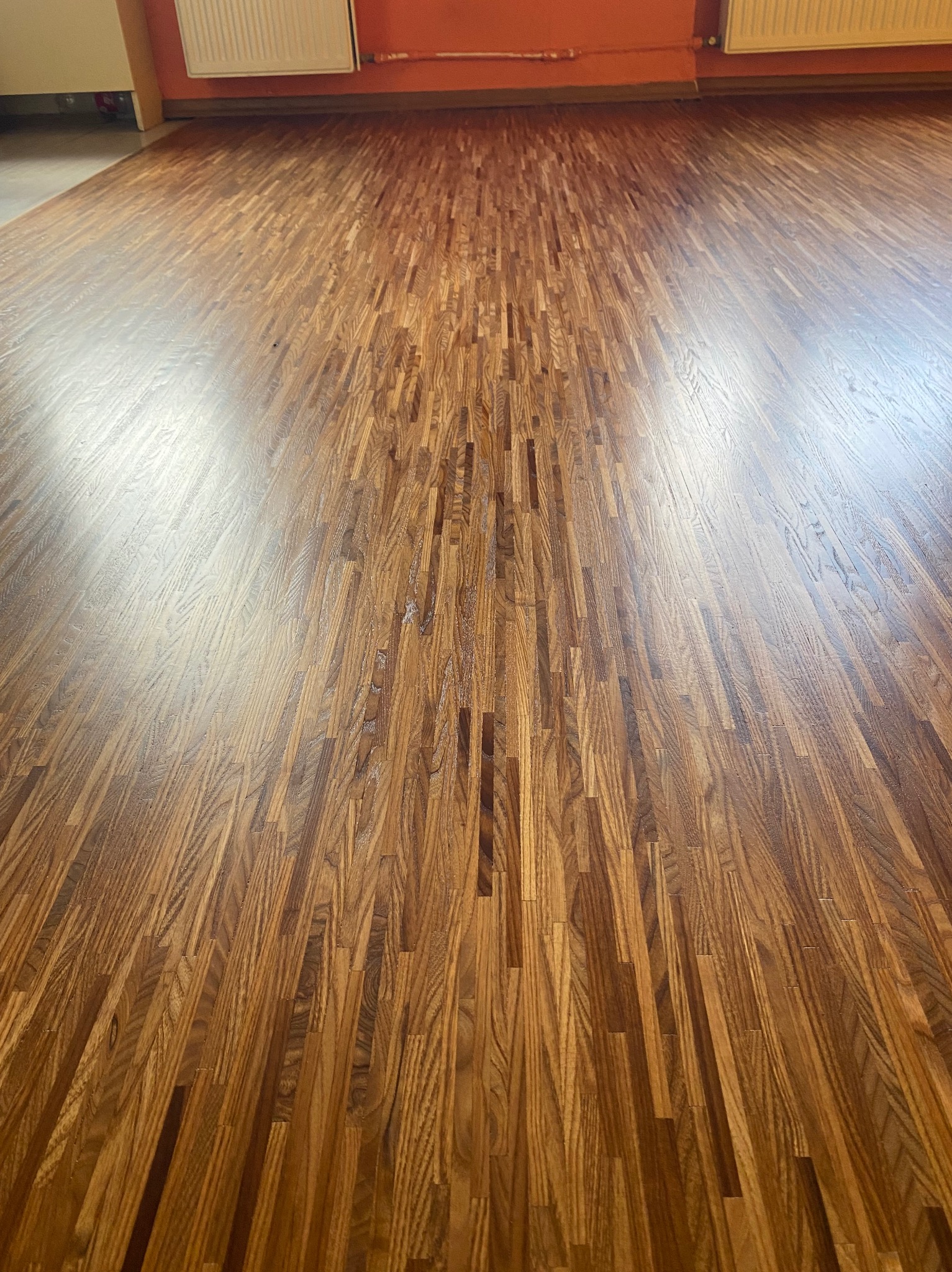 Parkiet z drewna dębowego ułożony w jodełkę, widoczne odbicia światła, pomarańczowa ściana i biały grzejnik w tle.