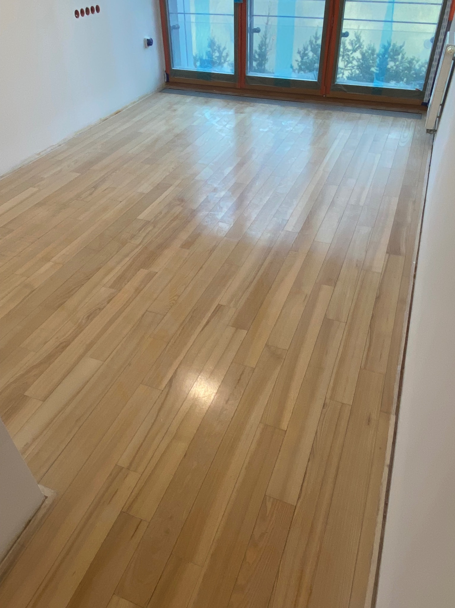 Świeżo wycyklinowana, jasna podłoga parkietowa w pustym pokoju, z widocznymi śladami po szlifowaniu i drobnymi niedoskonałościami, odbijająca światło z okna.