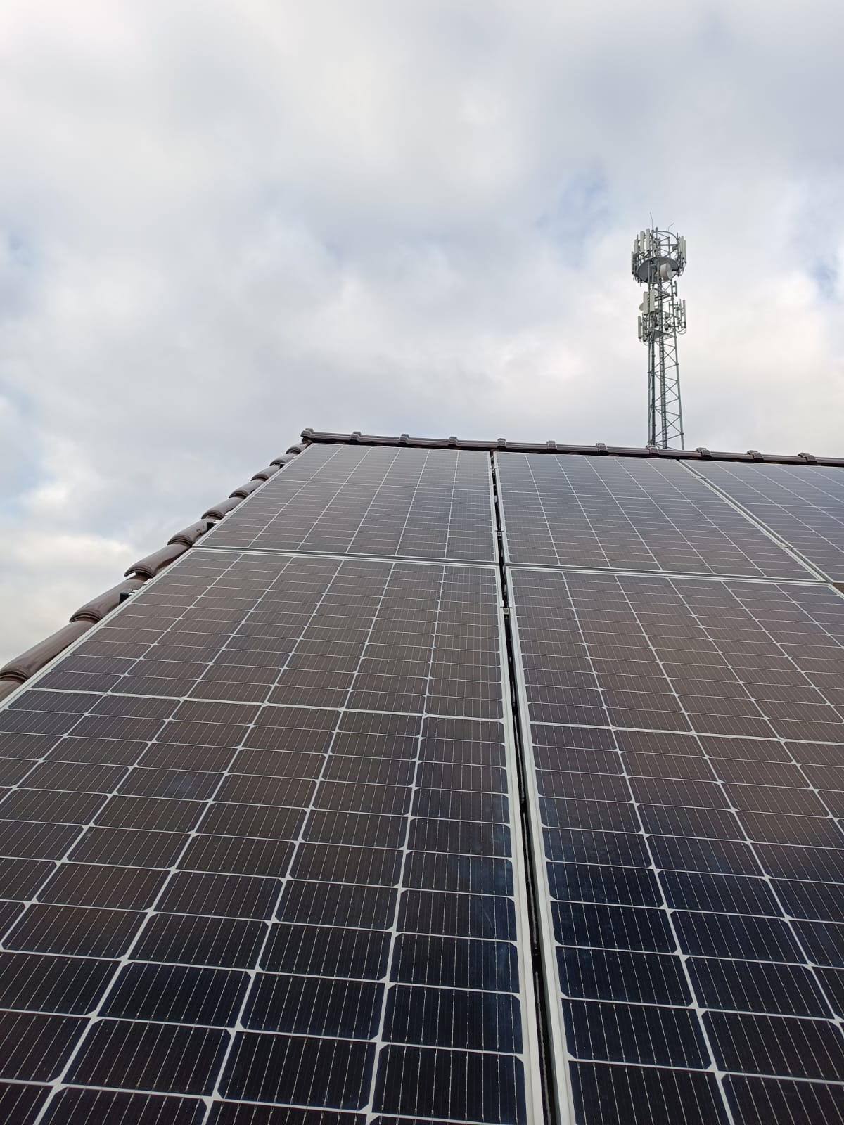 Panele fotowoltaiczne na brązowym dachu z dachówki ceramicznej, w tle wieża telekomunikacyjna i pochmurne niebo.
