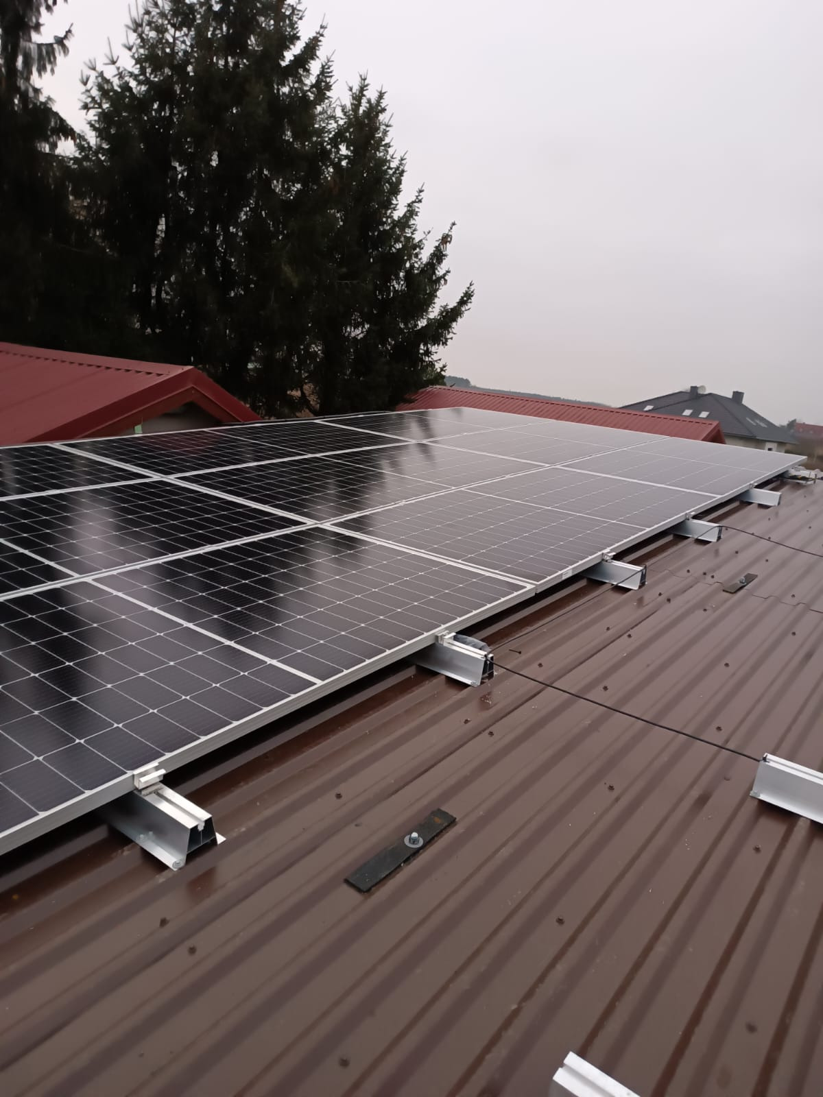 Panele słoneczne zamontowane na brązowym, blaszanym dachu w pochmurny dzień. Widoczne elementy montażowe i fragmenty innych dachów w tle.