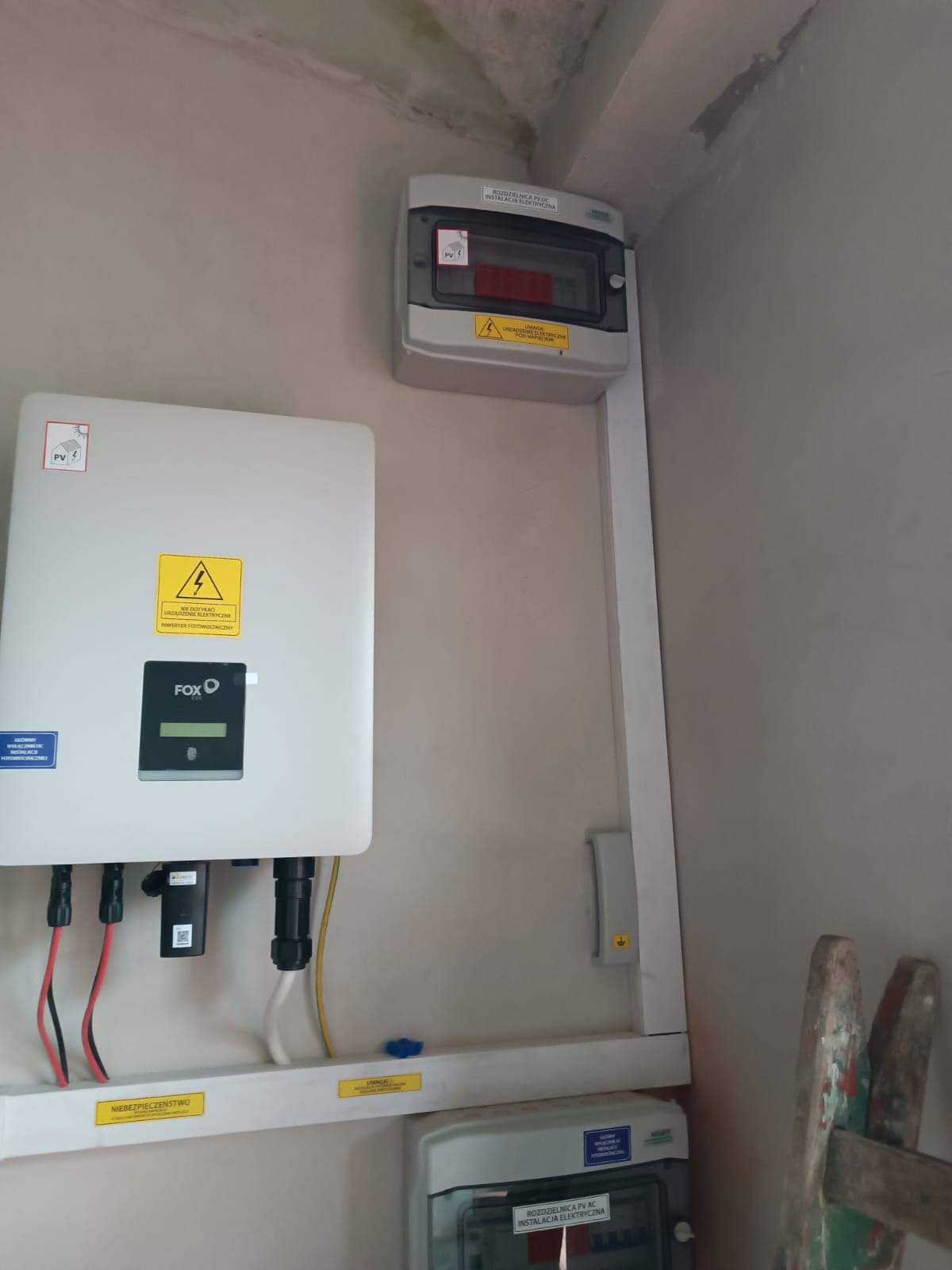 Zamontowany inwerter fotowoltaiczny marki FOXO z podłączonymi przewodami, obok rozdzielnica elektryczna PV i listwa z oznaczeniami ostrzegawczymi, widoczne elementy instalacji wewnątrz budynku.