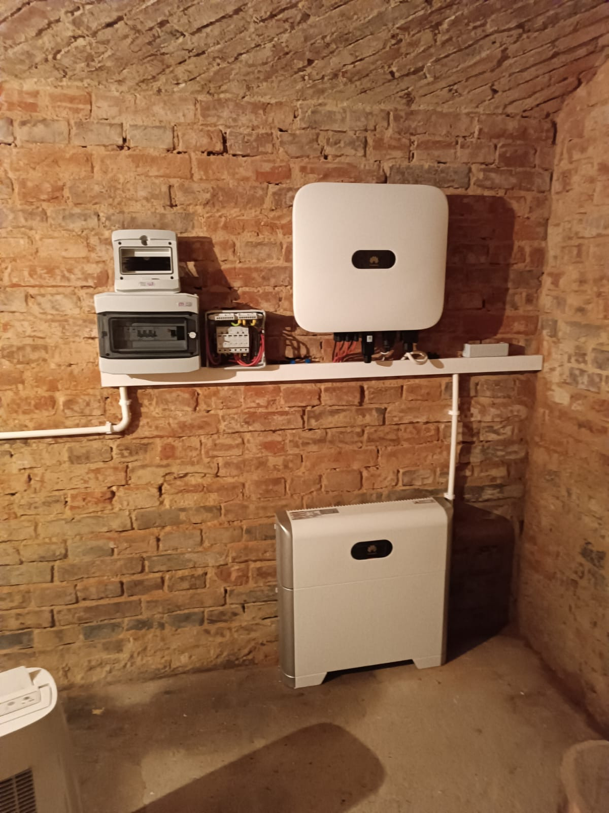 Instalacja wewnętrzna: biały inwerter i rozdzielnica na tle ceglanej ściany w piwnicy z łukowym sklepieniem.