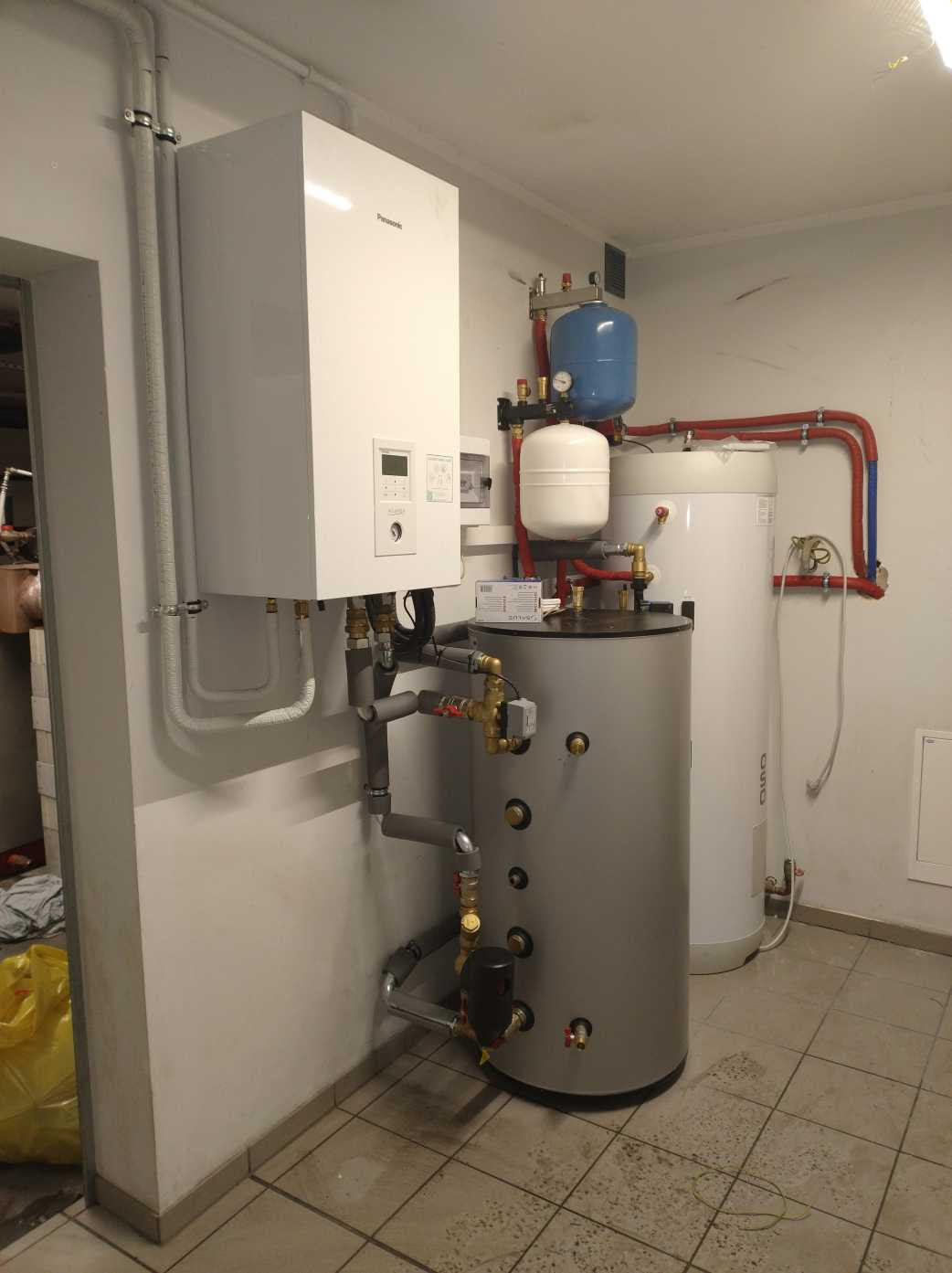 Zestaw dwóch zbiorników akumulacyjnych i wiszącego kotła gazowego Panasonic, połączonych rurami z zaworami, zainstalowany w pomieszczeniu z kafelkami na podłodze.