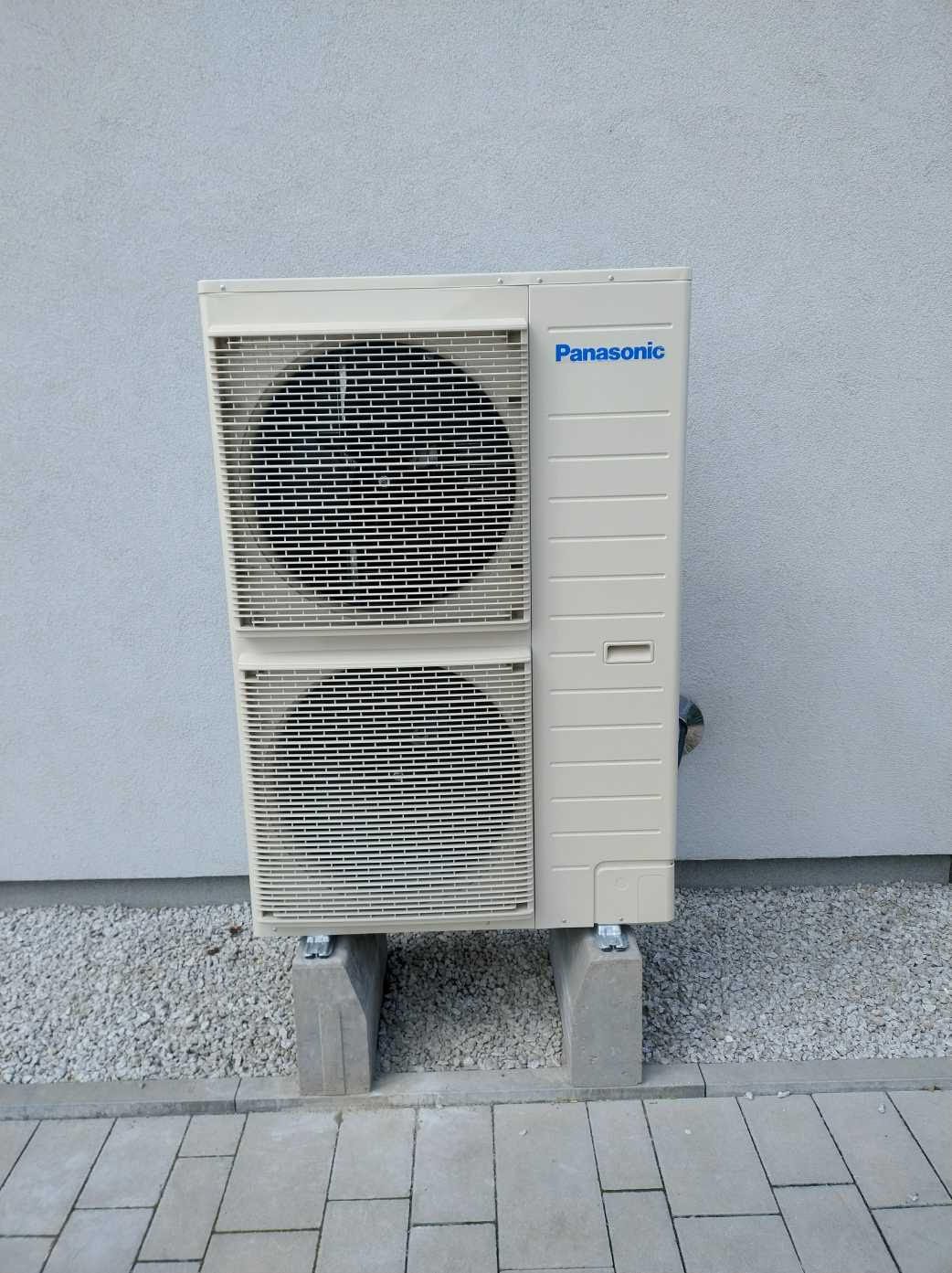 Kompaktowa, beżowa pompa ciepła Panasonic, umieszczona na betonowych podstawach przy jasnej ścianie budynku, otoczona drobnym żwirem i kostką brukową.