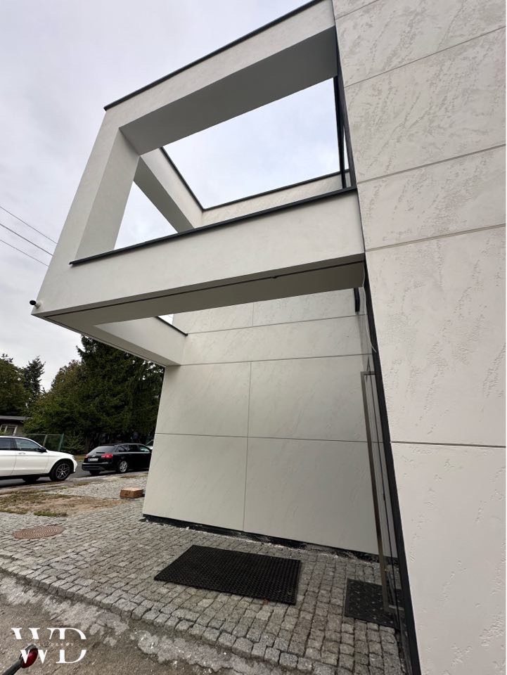 Nowoczesny budynek z jasną elewacją o fakturze, z widoczną konstrukcją w kształcie ramy nad wejściem, kostka brukowa na podjeździe.