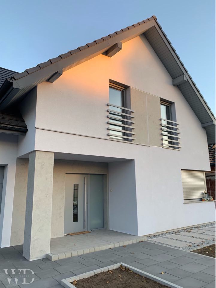 Nowoczesny dom jednorodzinny z jasną elewacją, szarą stolarką okienną i drzwiami wejściowymi, oświetlony popołudniowym słońcem, z widoczną strukturą tynku na kolumnach wejściowych.