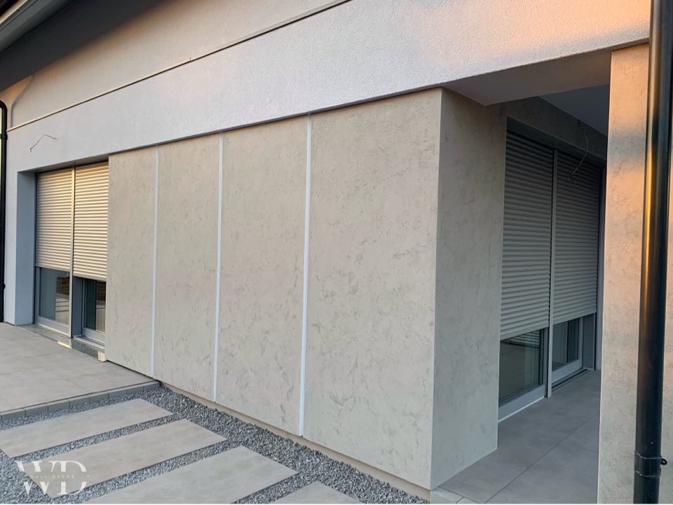 Elewacja budynku z tynkiem dekoracyjnym w odcieniach beżu z pionowymi paskami, widoczne okno z roletą zewnętrzną i fragment chodnika z płyt.