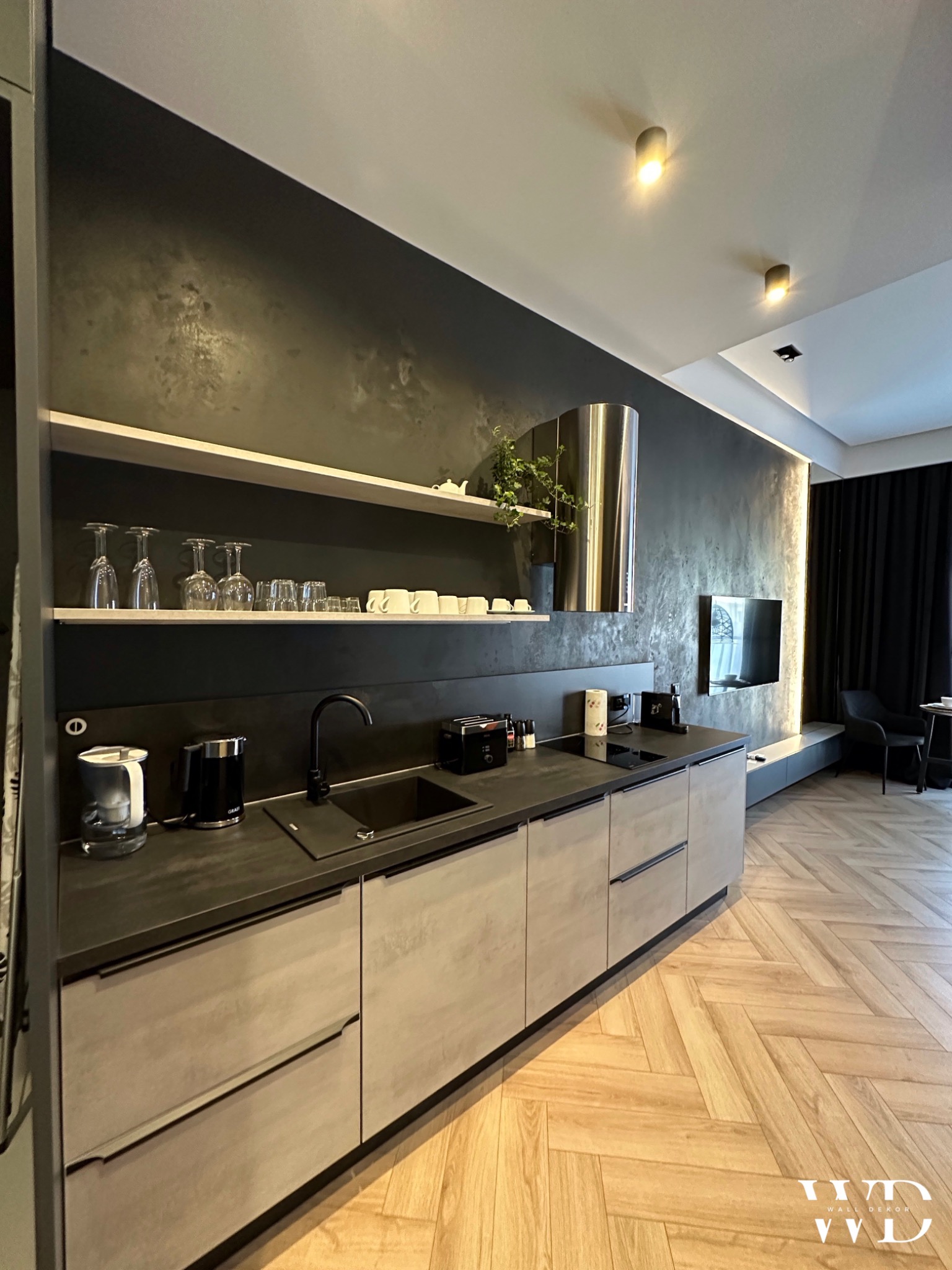 Nowoczesna kuchnia z ciemnoszarą ścianą o fakturze betonu, jasnymi szafkami, drewnianą podłogą w jodełkę i telewizorem w tle.