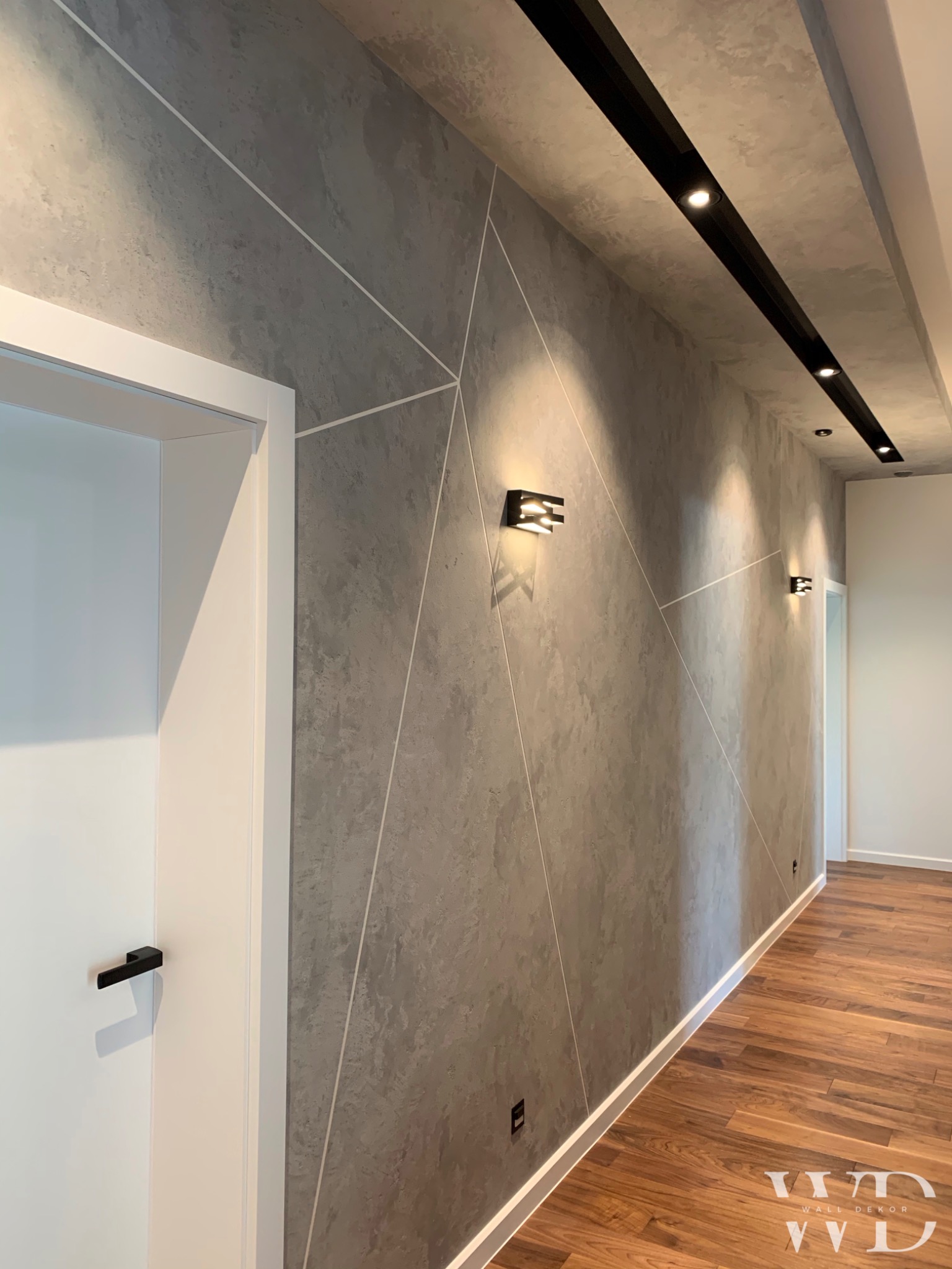 Szara ściana z efektem betonu, zdobiona białymi geometrycznymi liniami, oświetlona nowoczesnymi kinkietami i listwą sufitową z reflektorami, wykończona drewnianą podłogą.