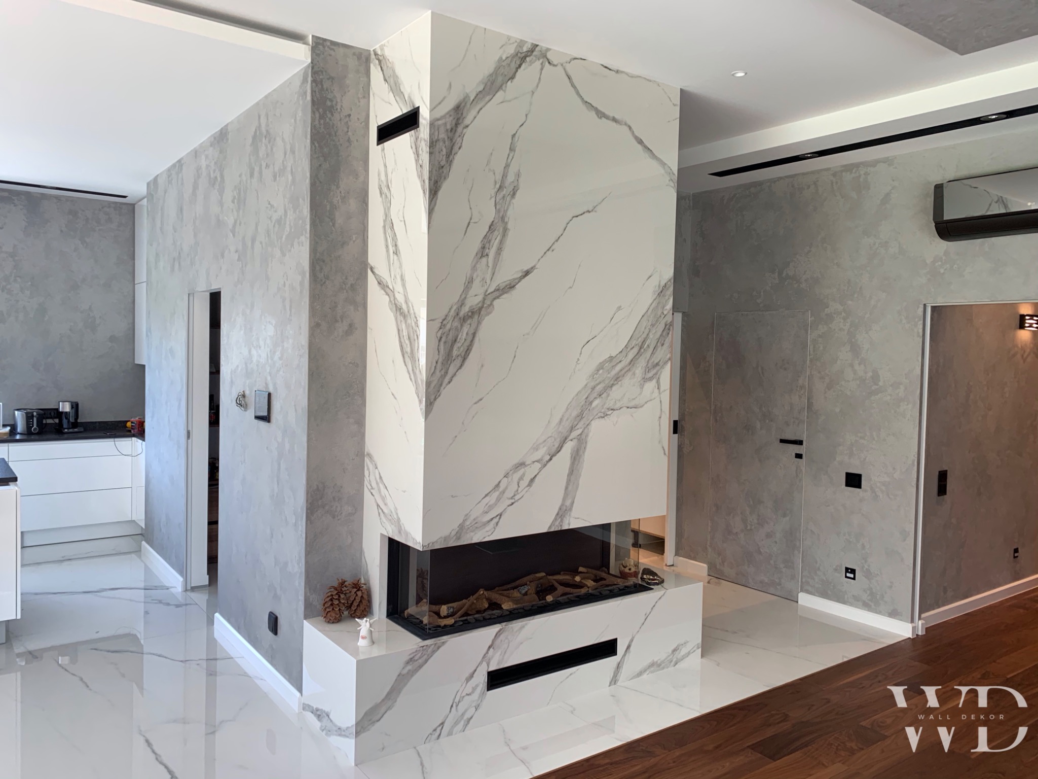 Salon z nowoczesnym kominkiem obłożonym płytami imitującymi marmur, szare ściany wykończone dekoracyjnym tynkiem, białe podłogi z żyłkowaniem, widoczny fragment kuchni i parkietu.