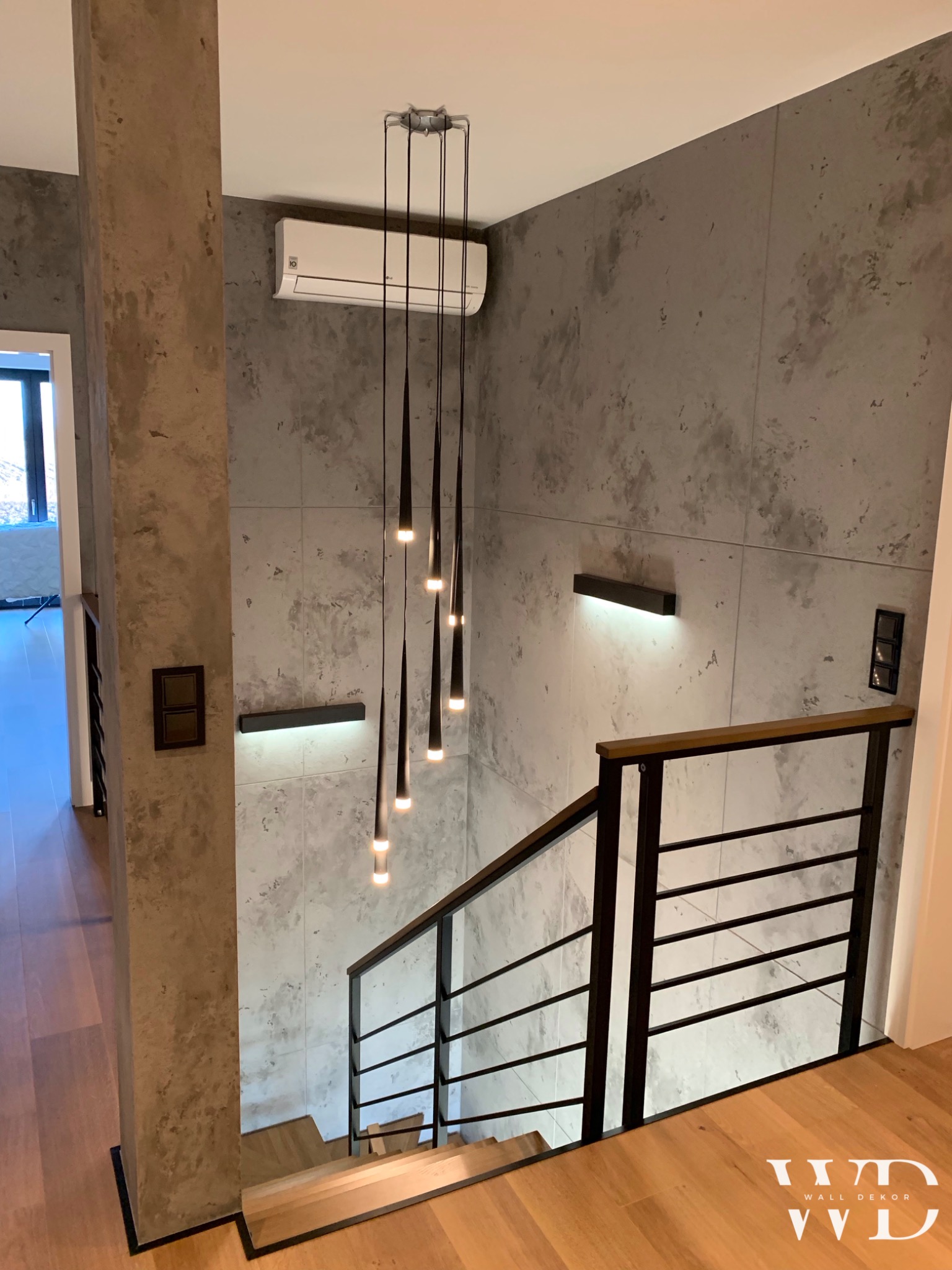 Klatka schodowa z szarą ścianą o fakturze betonu, oświetlona wiszącymi lampami w kształcie sopli i podłużnymi kinkietami, z widocznym klimatyzatorem LG na ścianie.